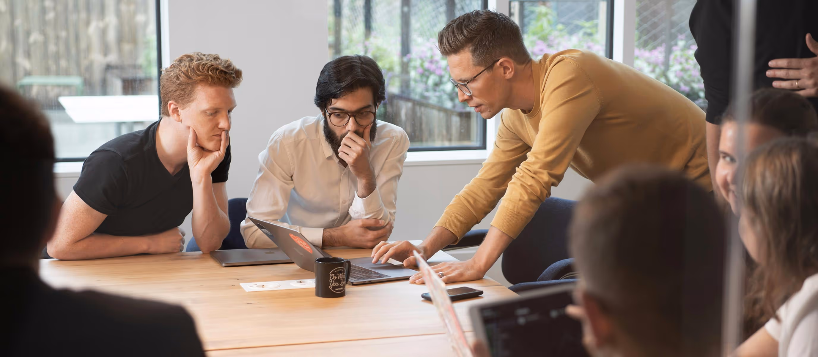 Three men in a meeting room focused on a laptop screen, with one standing and pointing at the laptop keyboard while two others sit and watch attentively.