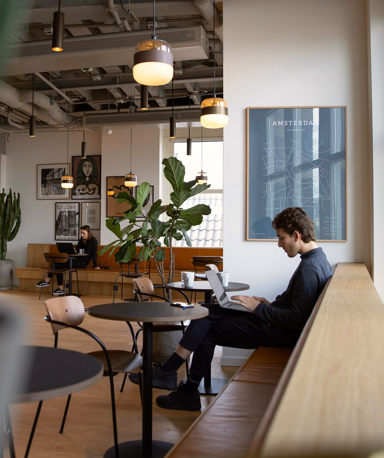 Two people working on laptops in a modern cafe with hanging lights, potted plants, and Amsterdam map artwork.