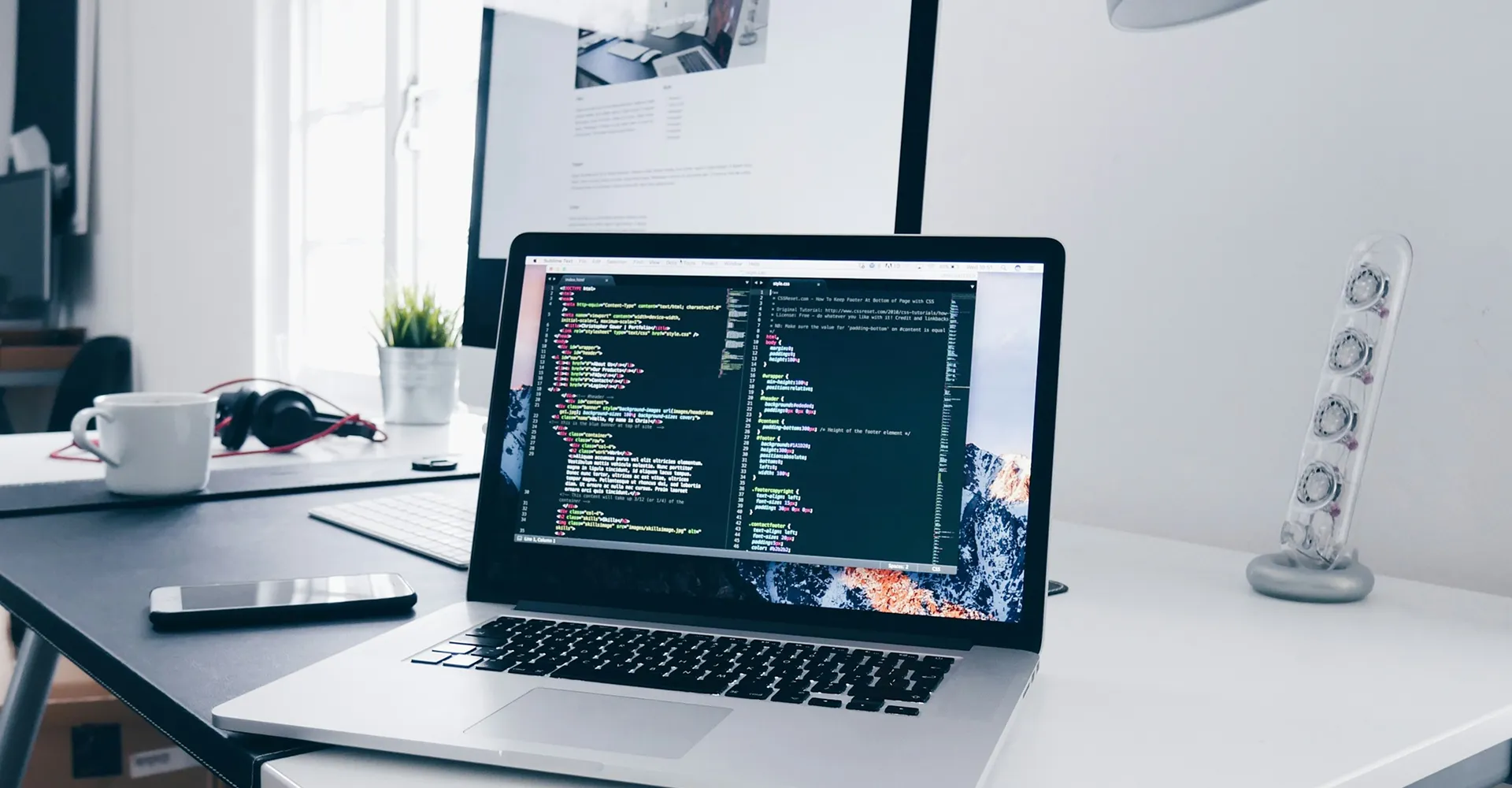 Laptop on desk displaying code editor with HTML and CSS code beside a smartphone and coffee cup.