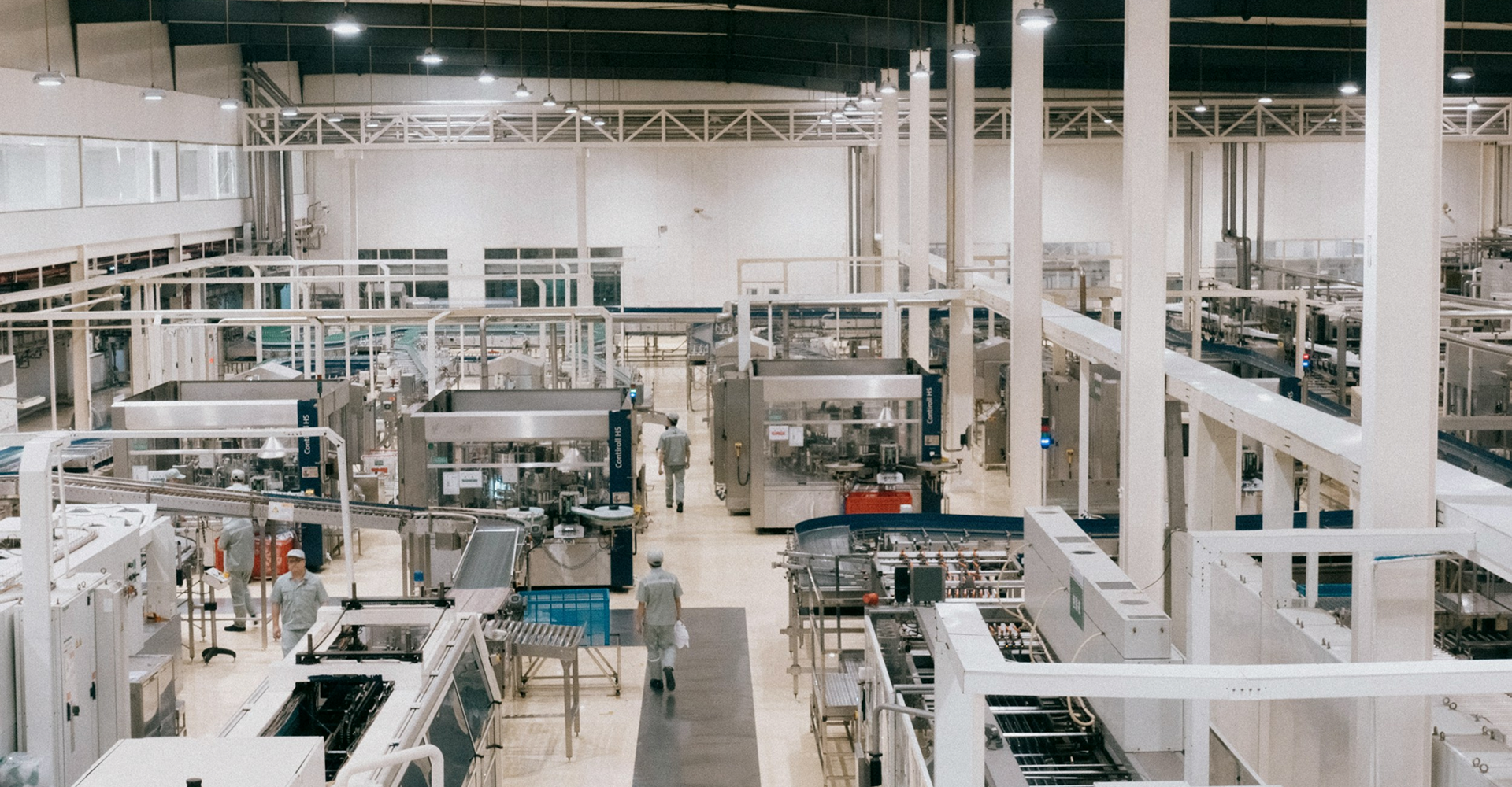 Workers in uniform walking among large automated machinery inside a bright, spacious factory.