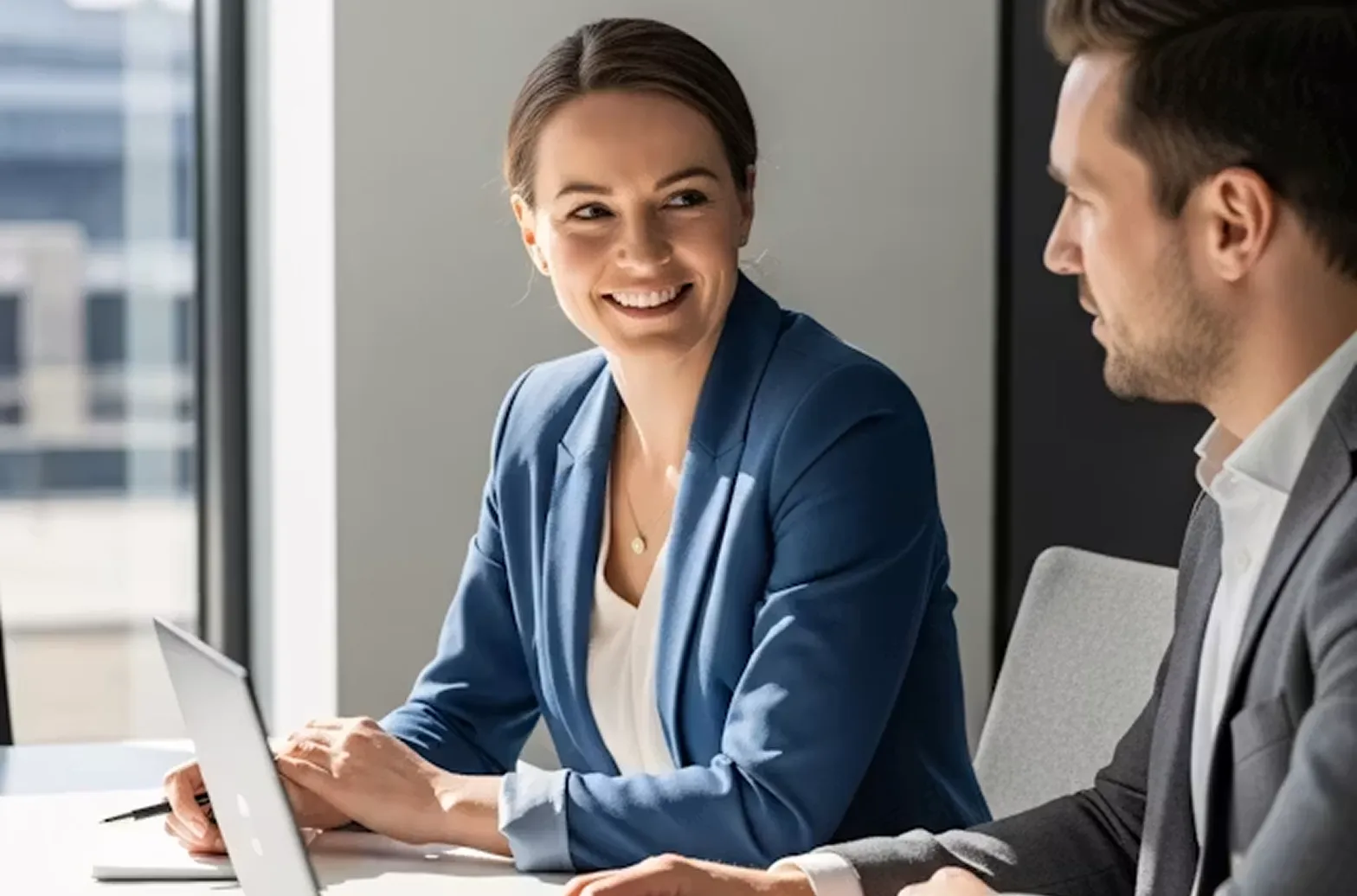 Smiling woman in blue blazer talks with man in gray suit at a desk with a laptop.
