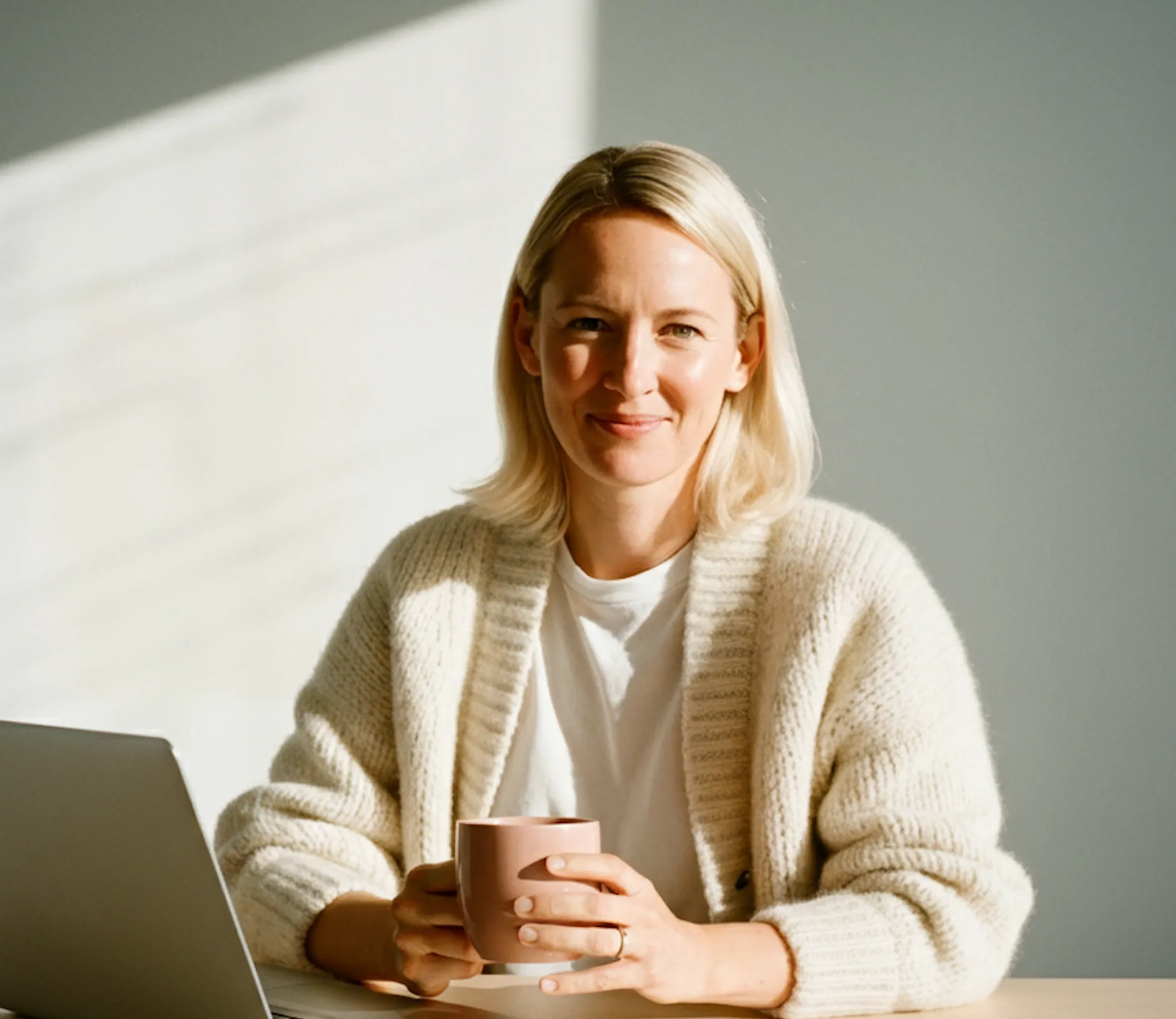 Blonde woman in a white sweater holding a mug, sitting at a table with a laptop in soft light.