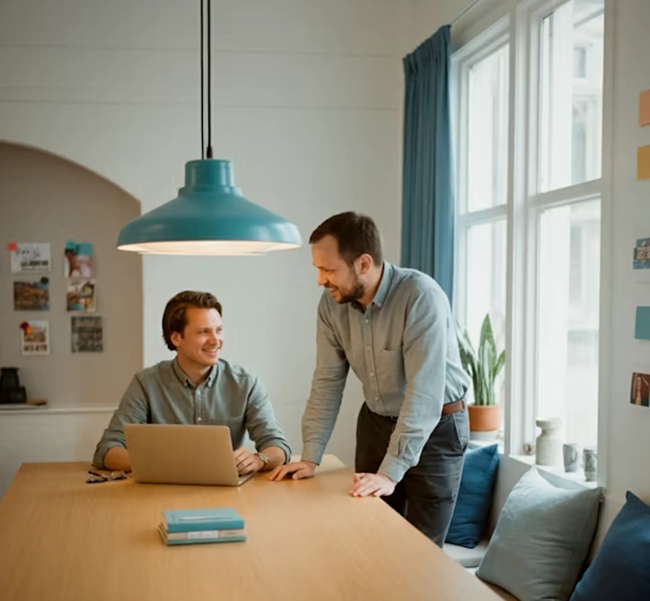 Two men smiling and working together at a wooden table with a laptop in a bright office.
