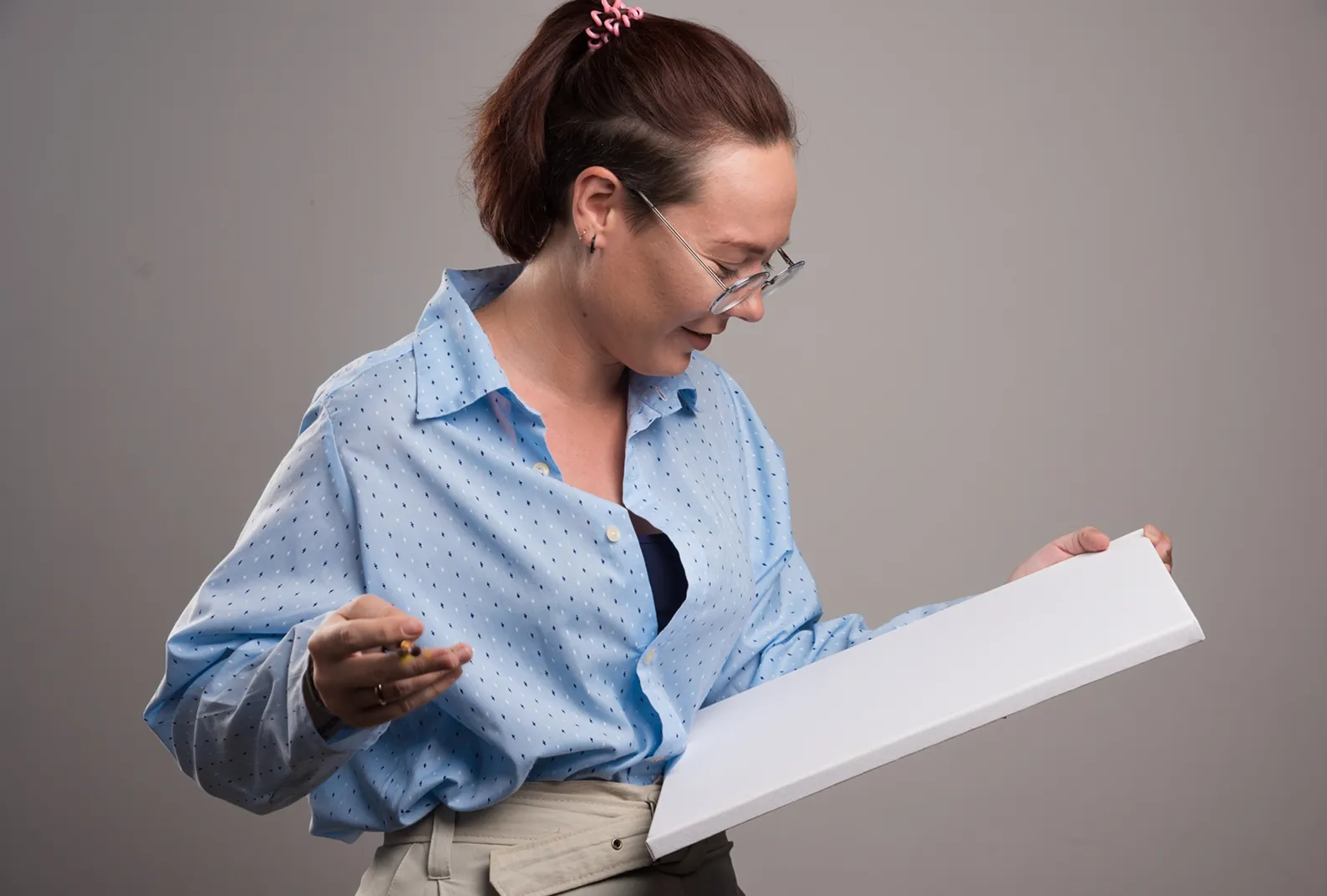 Woman with glasses holds a blank canvas and a pencil, looking at the canvas while smiling.