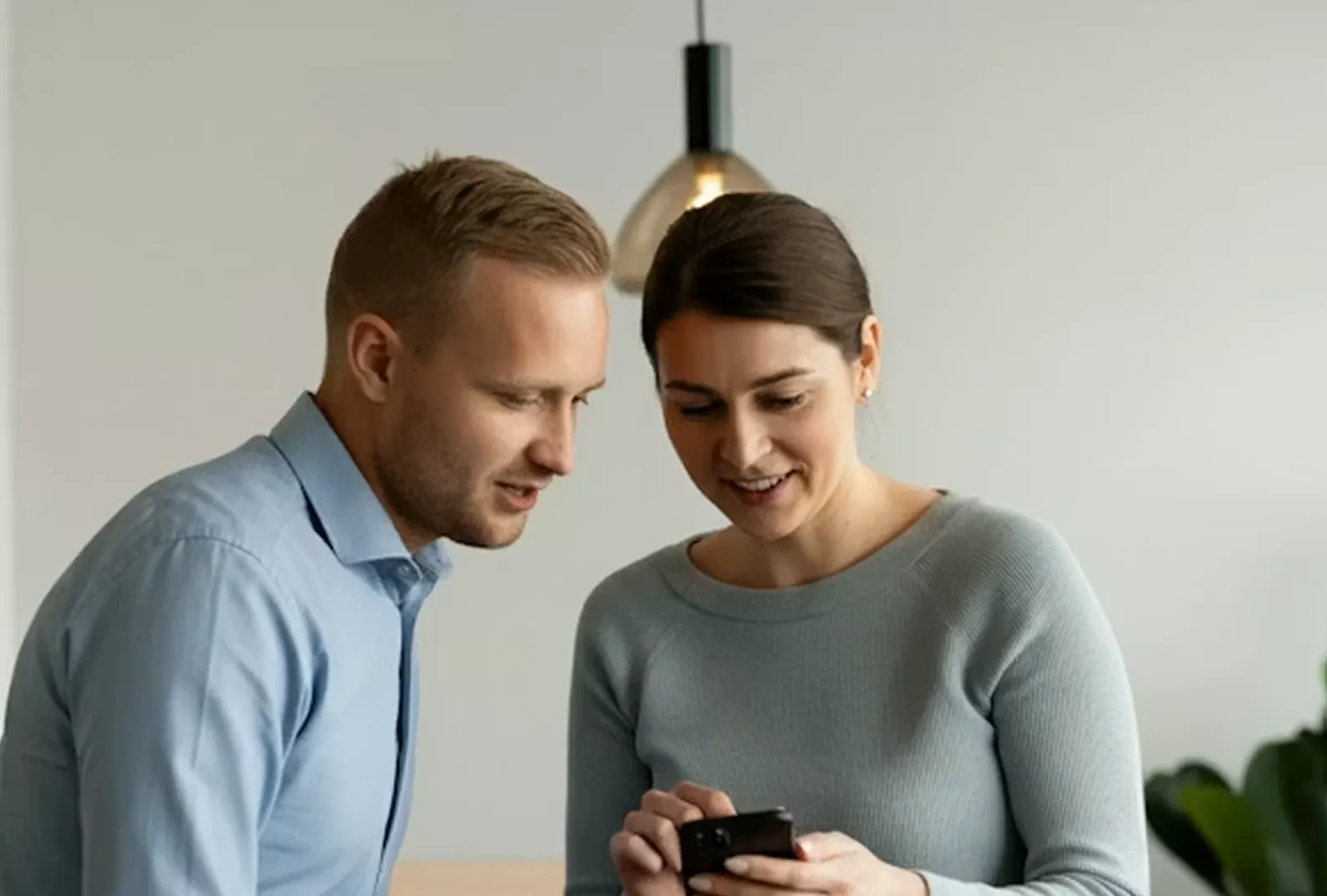 Man and woman looking at a smartphone together indoors with a hanging light fixture above.
