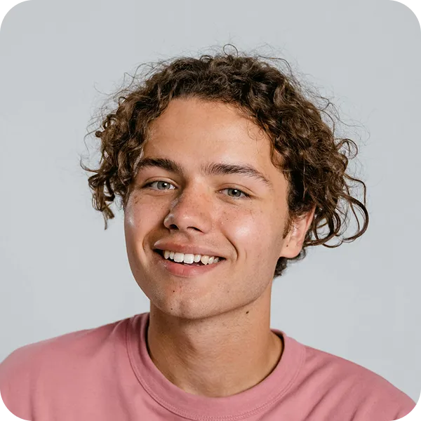 Smiling young man with curly hair wearing a pink shirt against a light gray background.