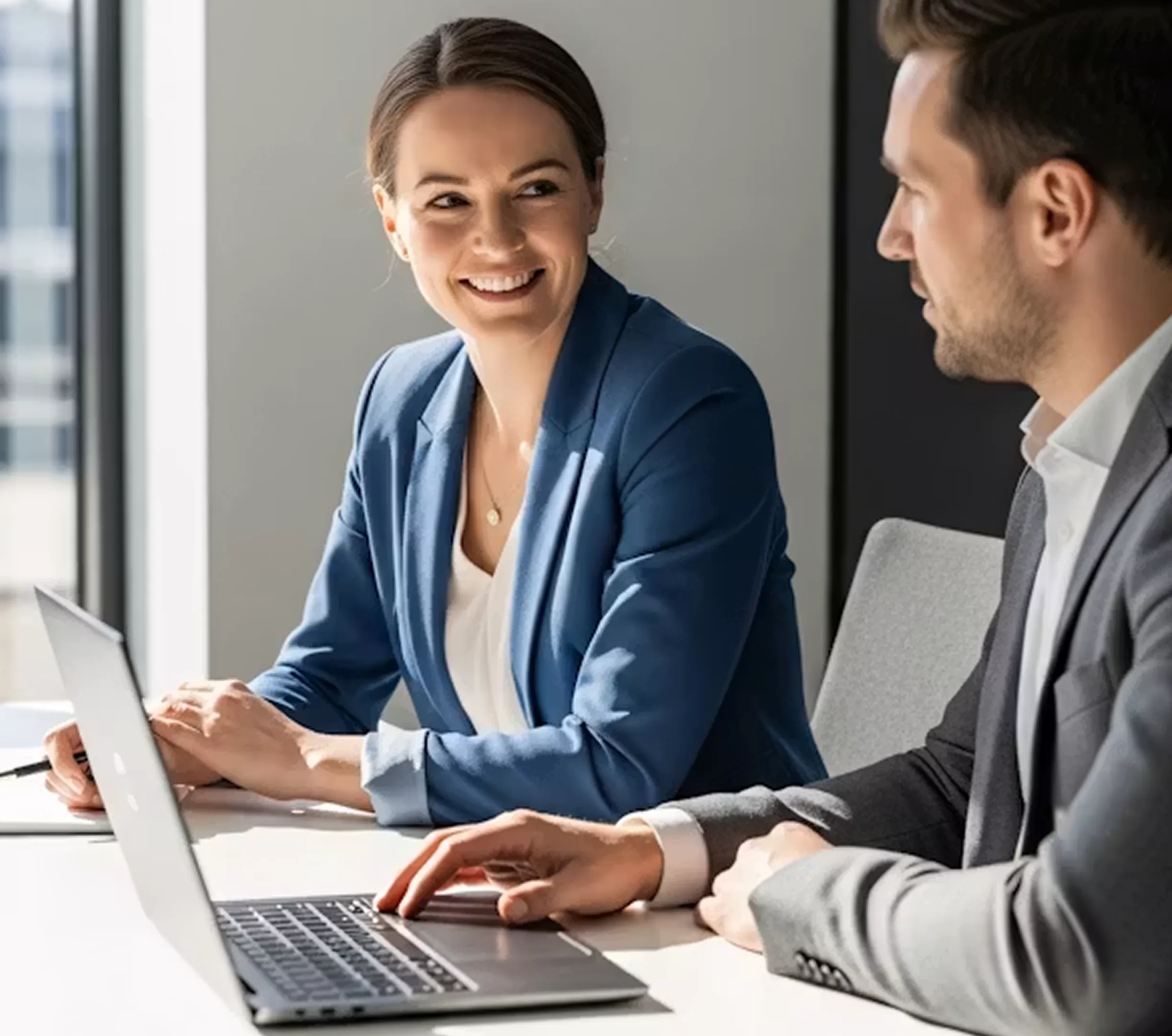 Woman in blue blazer smiling at man in gray suit working on a laptop in an office.
