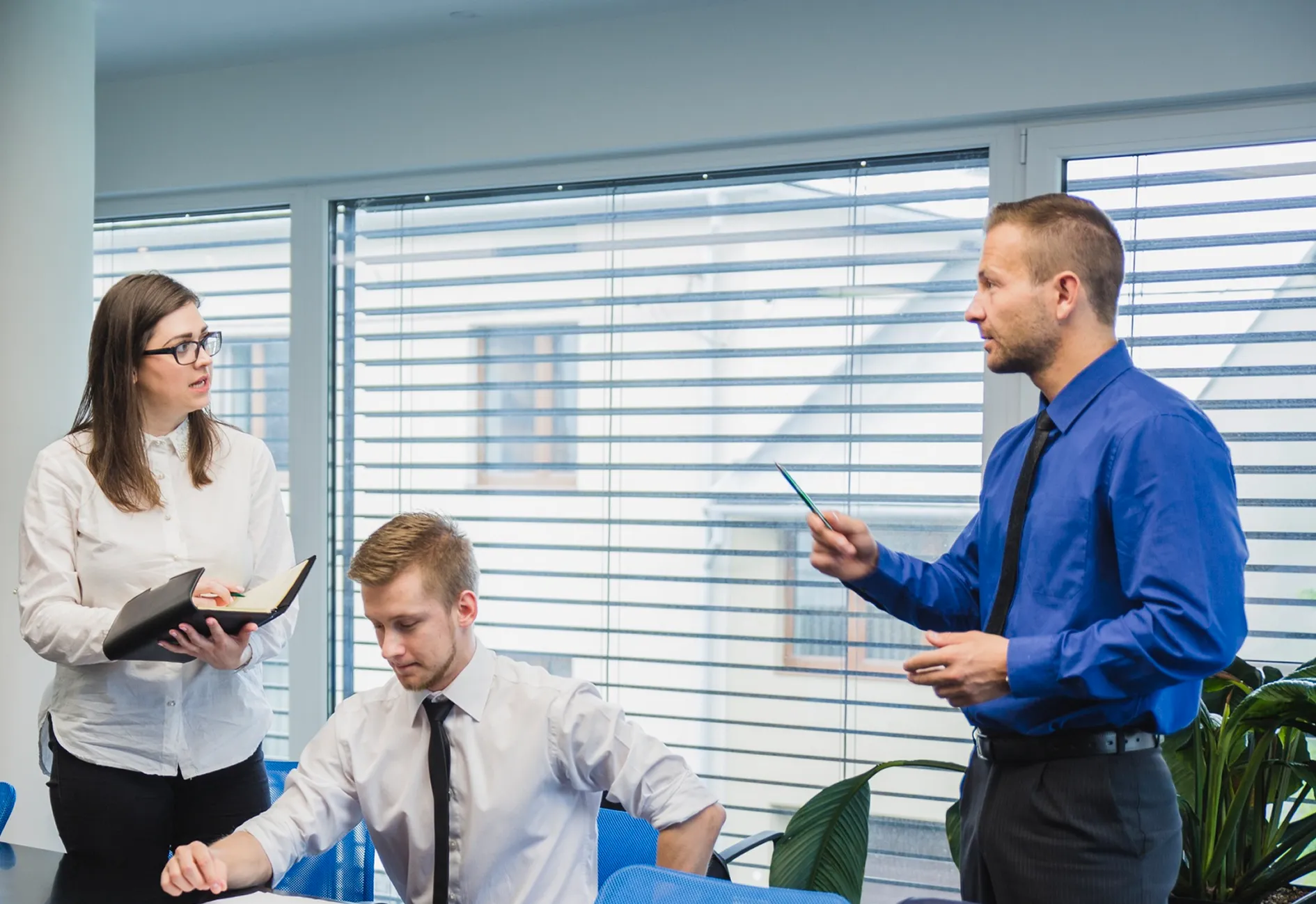 Three business people in office meeting: one standing woman with notebook, one seated man, one standing man speaking.