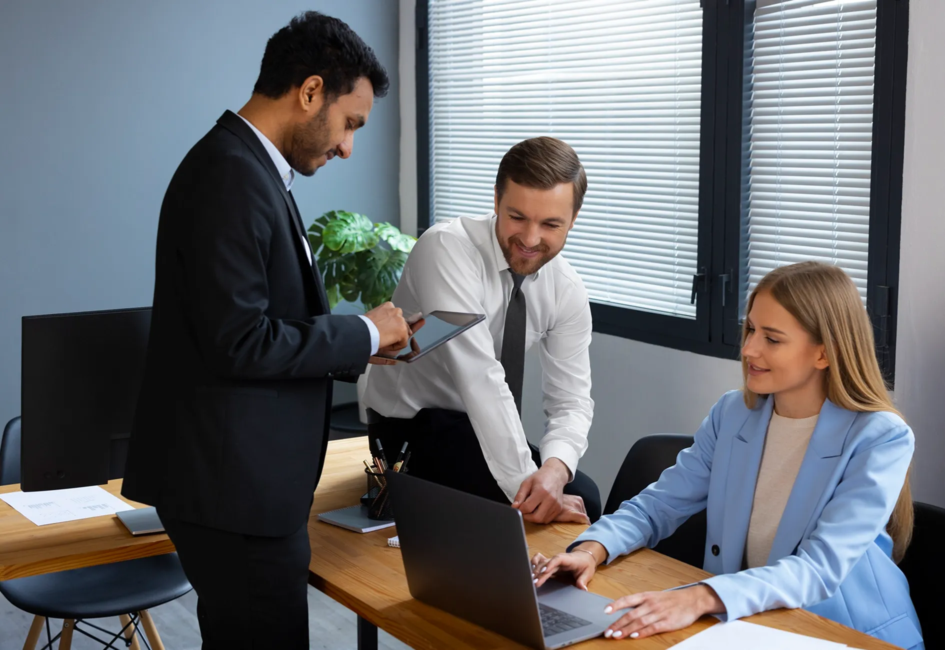 Three office workers in business wear collaborating at a desk with a laptop and tablet.