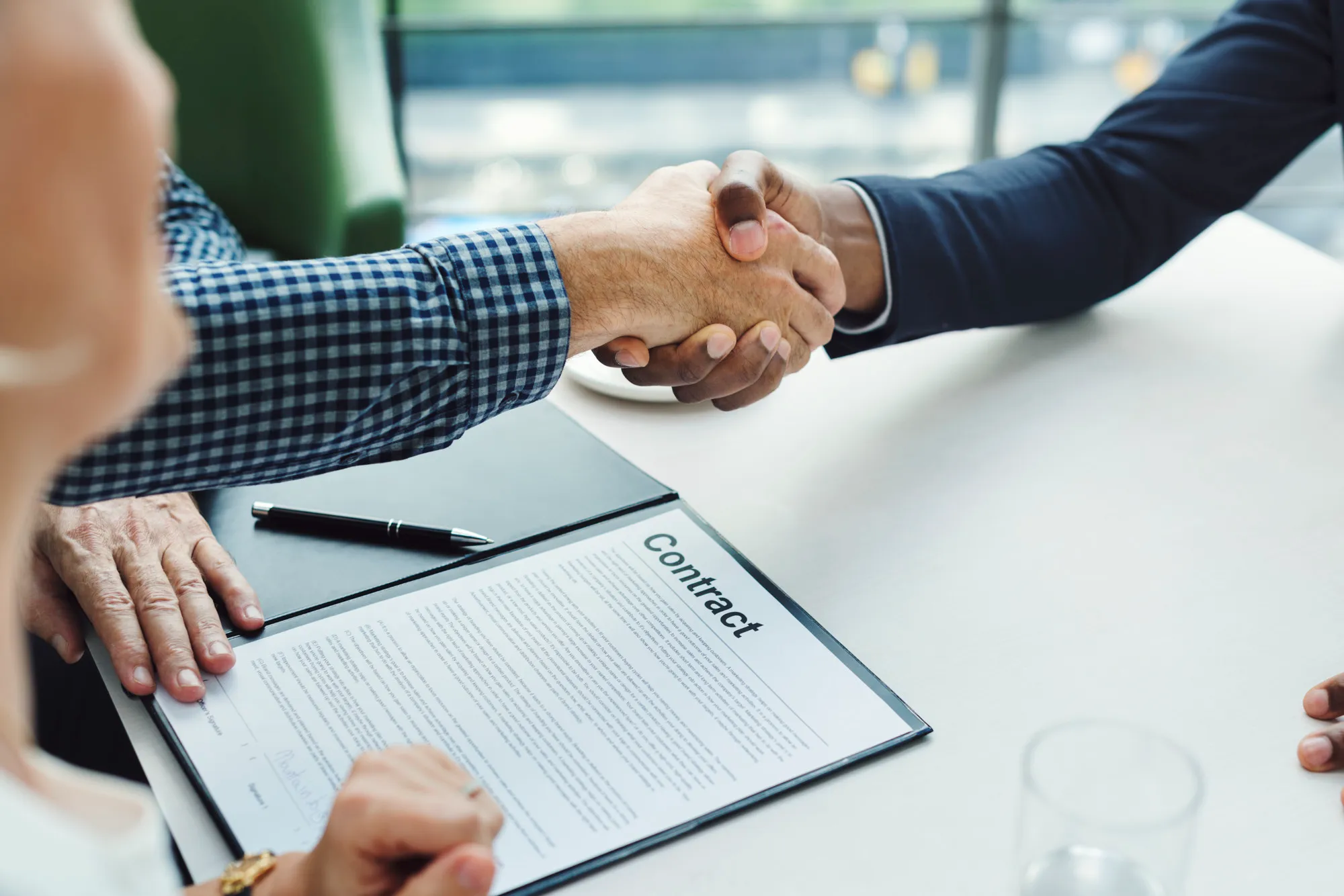 Two people shaking hands over a signed contract on a table with a pen nearby.