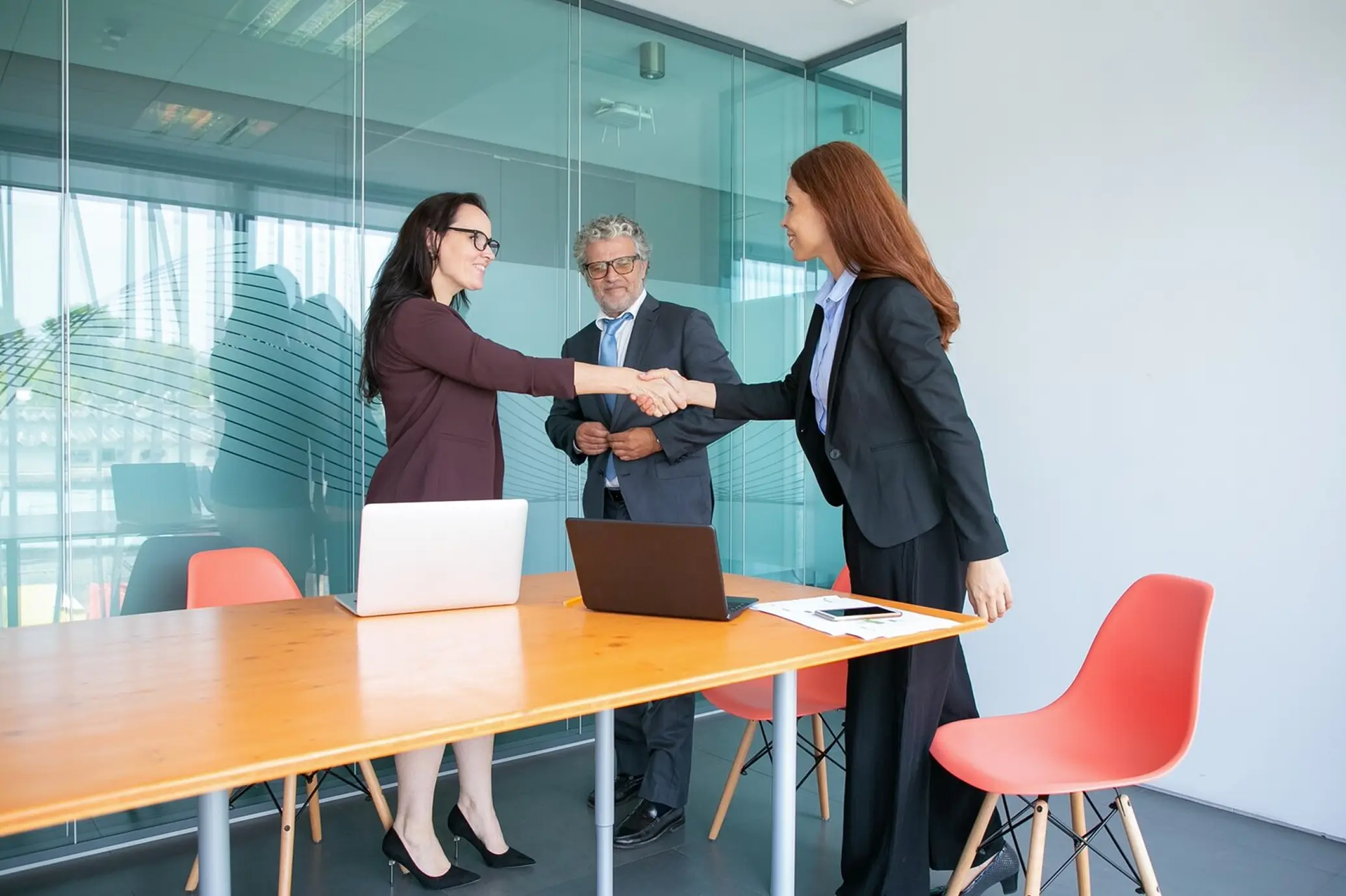 Two businesswomen shaking hands in an office meeting room with a man standing behind them.