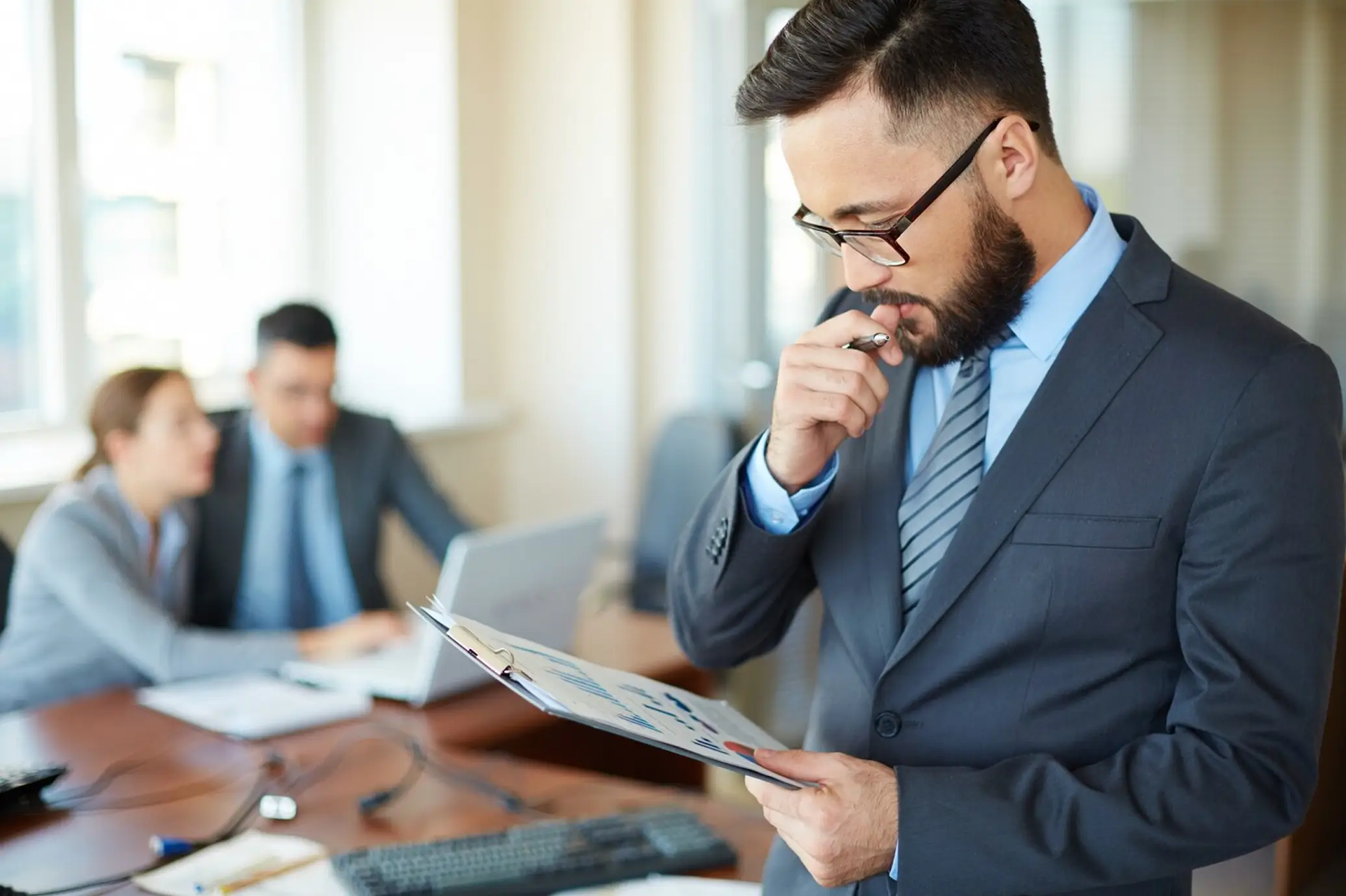 Man in suit and glasses thoughtfully reviewing a clipboard with charts in an office.