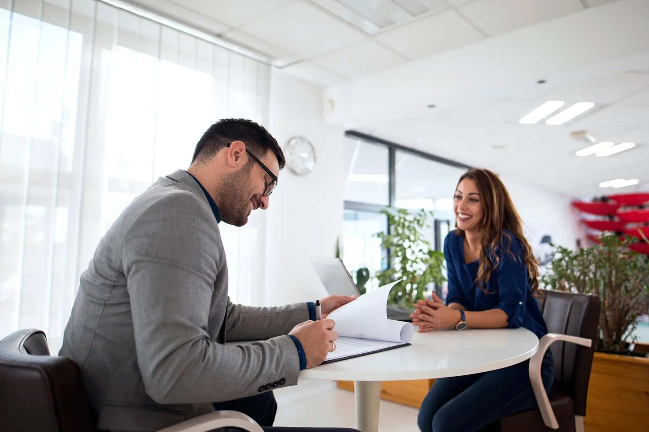 Man in gray suit reviewing documents while woman in blue shirt smiles during office meeting.