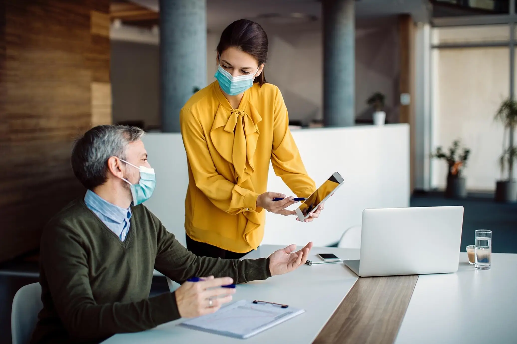 Two masked coworkers discussing while woman shows tablet screen to seated man in office.