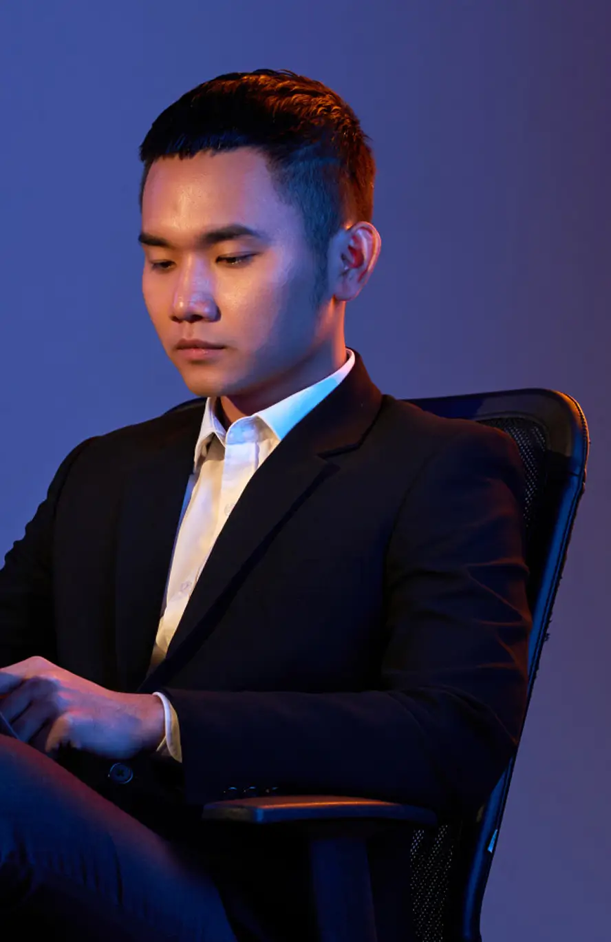 Young man in a suit sitting on a chair against a blue-purple blurred background.