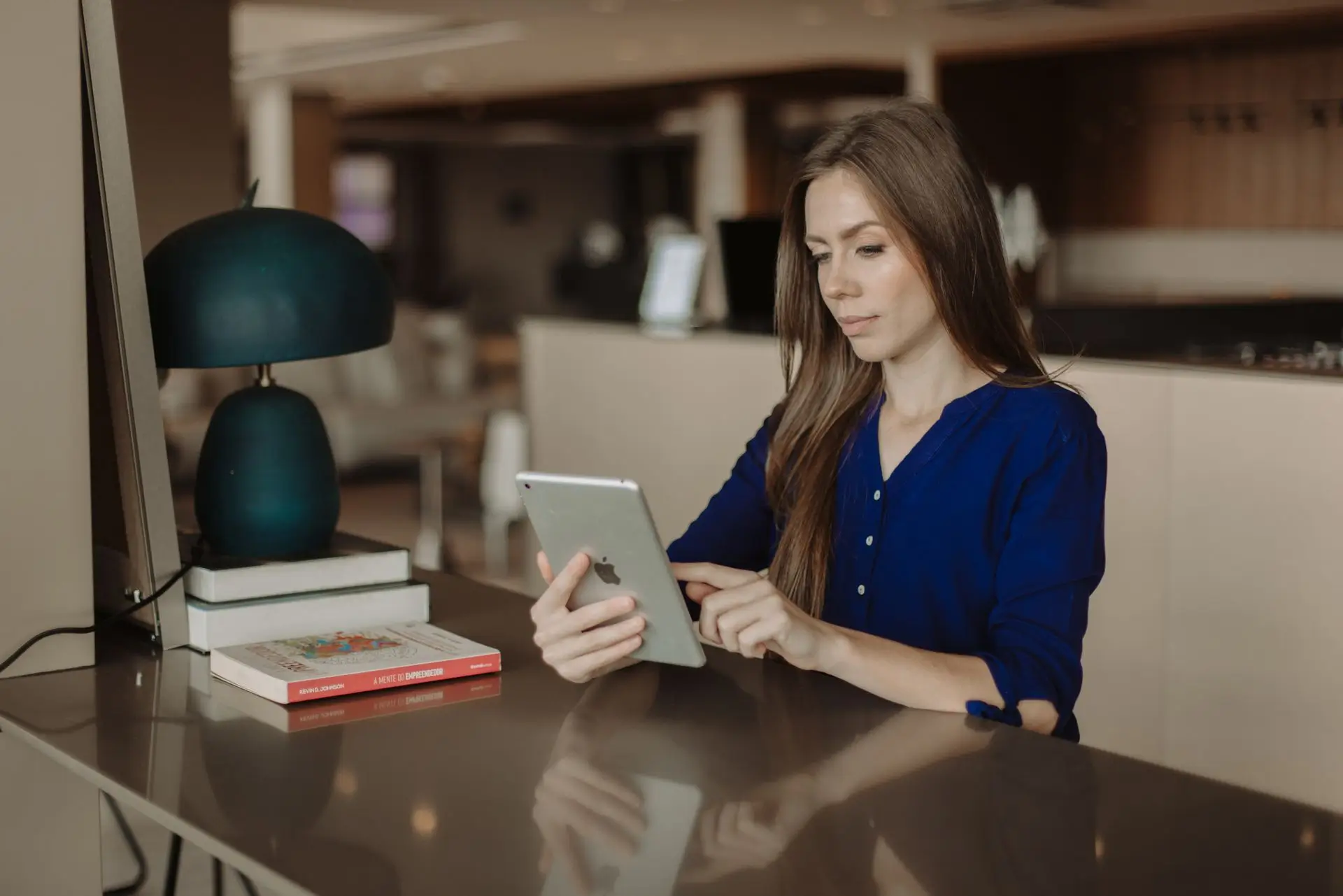 Woman in blue shirt sitting at reflective table using a tablet in a cozy room with a lamp and books.