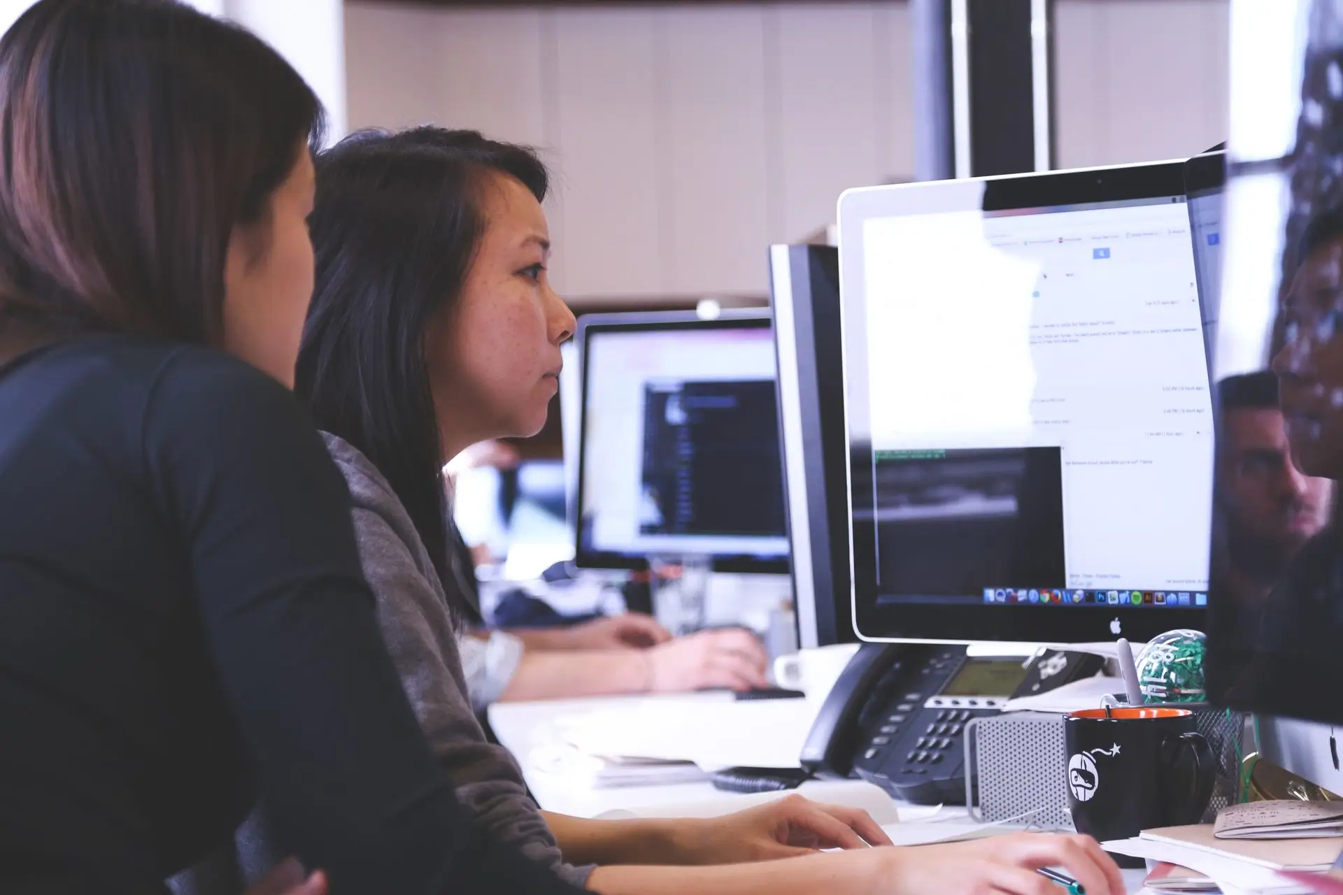 Two women working together at a computer in an office with monitors and office supplies on the desk.