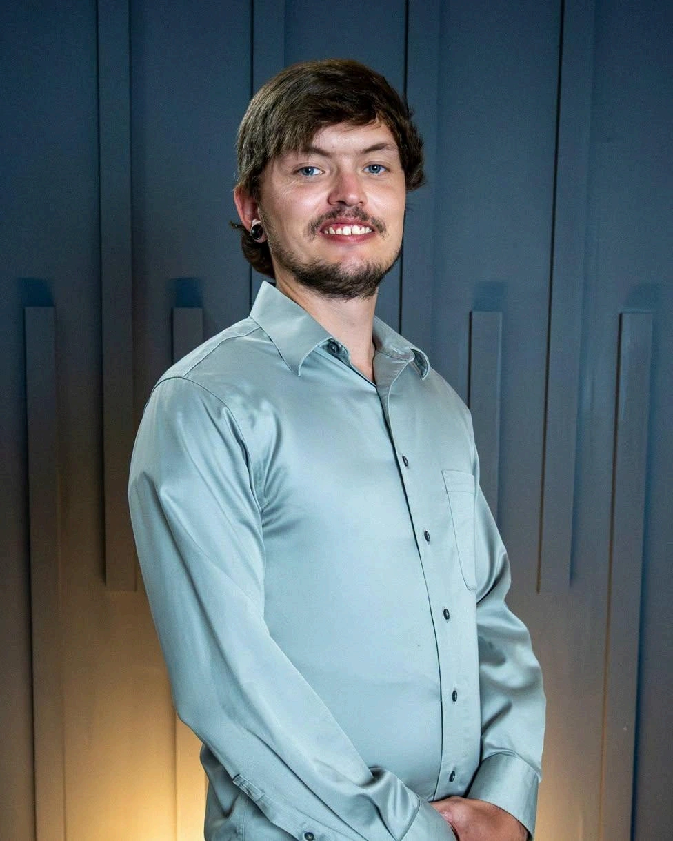 Smiling man with dark hair and beard wearing a light blue dress shirt against a dark paneled background.