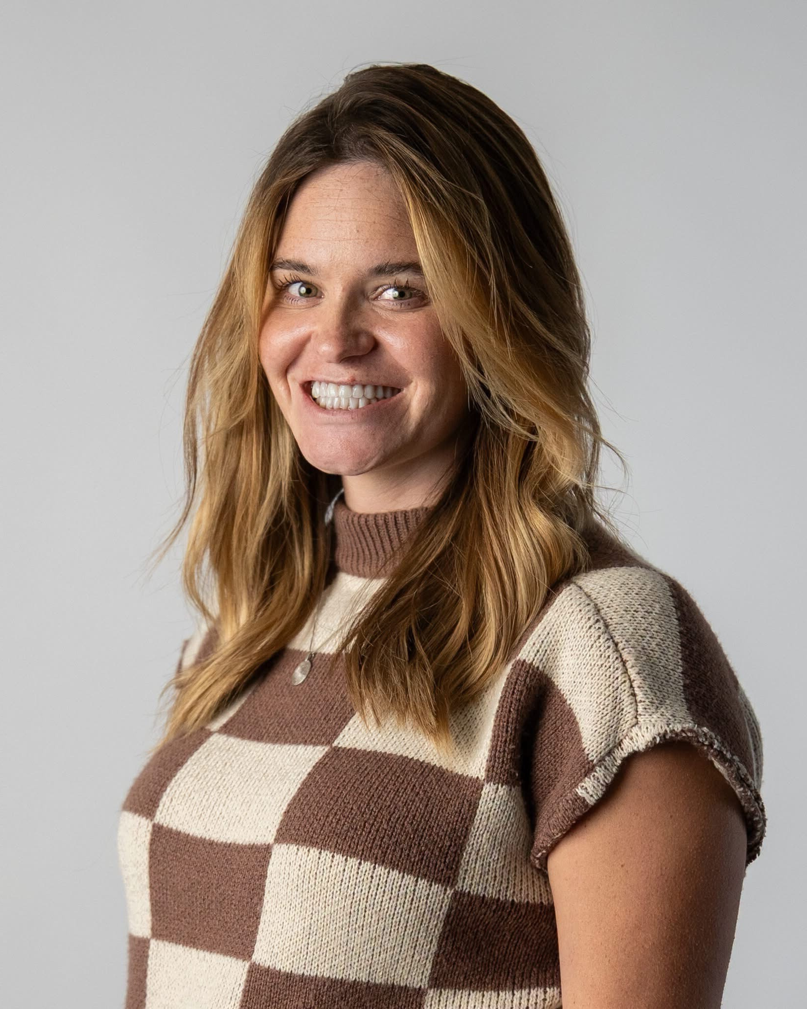 Smiling woman with long blonde hair in brown and cream checkered knit top against gray background.