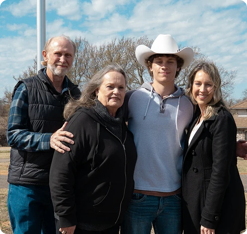 Four people smiling outdoors under blue sky, including young man in white cowboy hat and casual clothes.