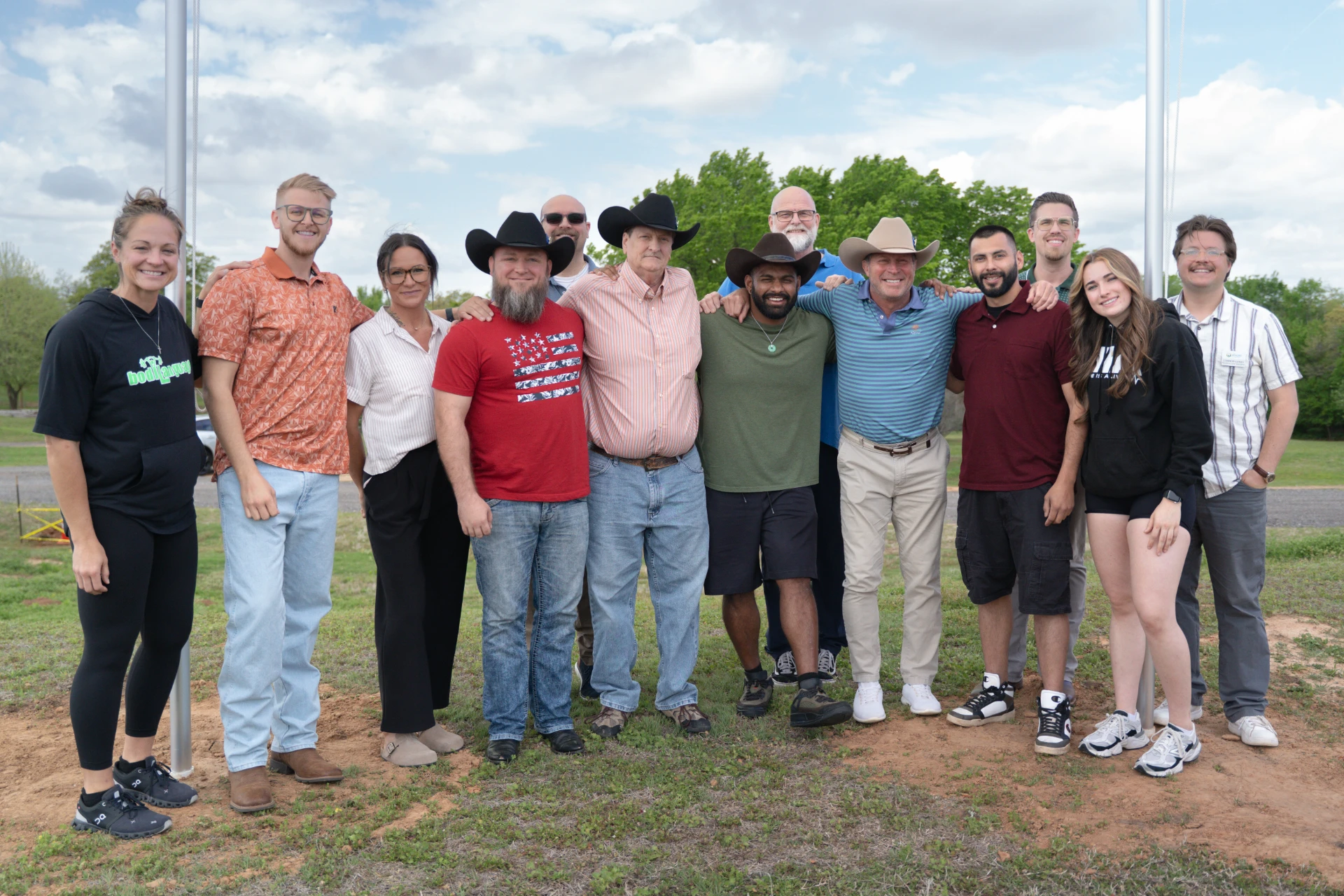Group of twelve adults outdoors standing close with arms around each other smiling under a partly cloudy sky.