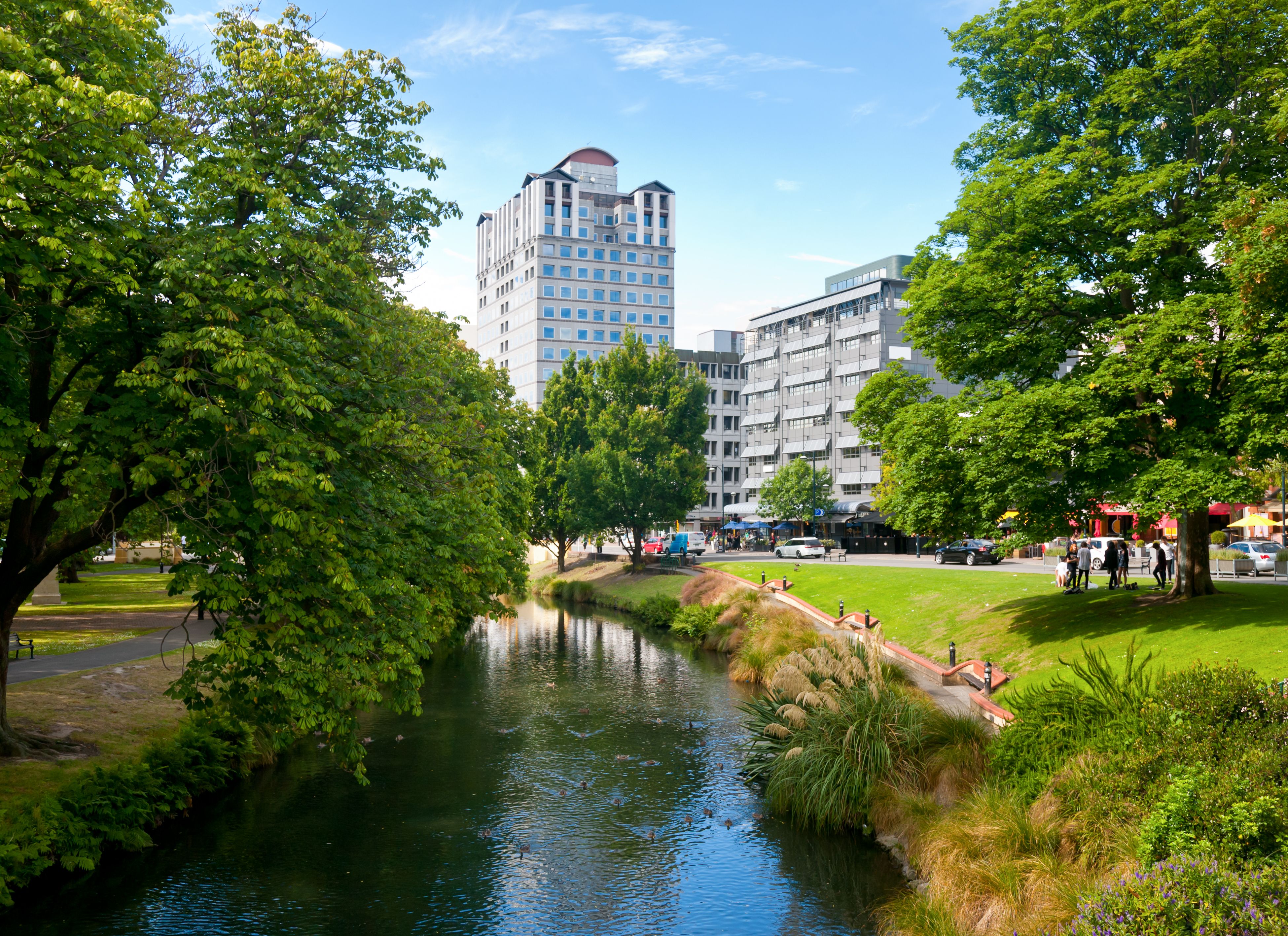Urban river flanked by green trees and grass, with buildings and people in the background on a sunny day.