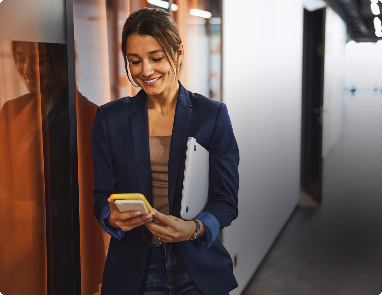 Une professionnelle consulte son téléphone, un ordinateur portable sous le bras, dans un couloir de bureaux