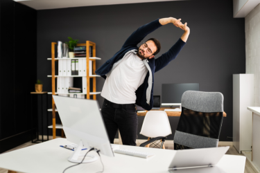A man stands beside his office desk and stretches sideways with both arms raised overhead.
