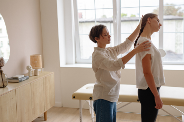 A practitioner gently positions a standing patient’s head and shoulder during a neck assessment in a treatment room.