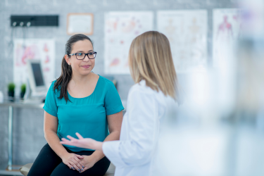 A patient wearing glasses sits on an examination table and talks with a healthcare professional in a white coat.