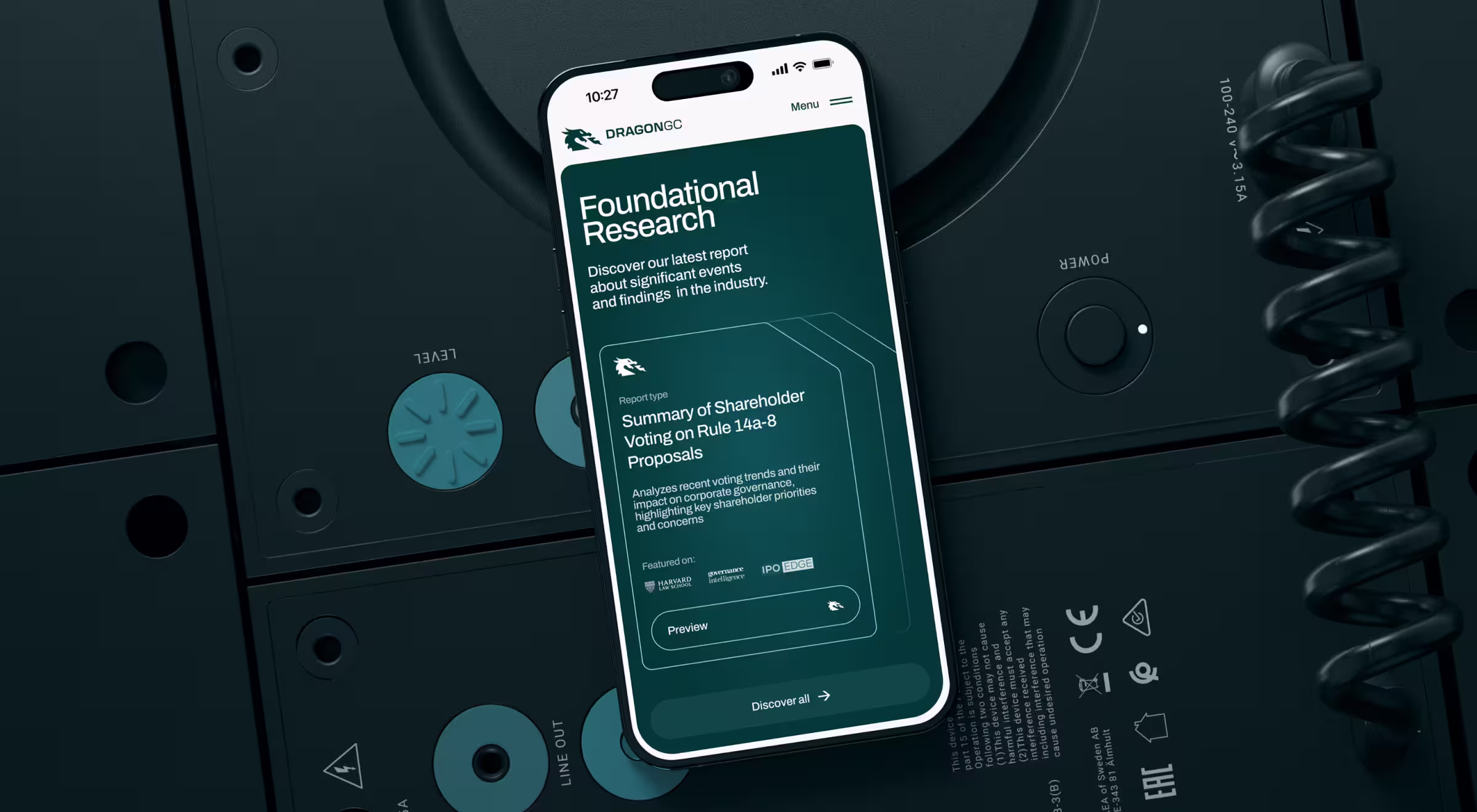 Smartphone displaying the DRAGONGC ‘Foundational Research’ report page, placed on top of dark industrial equipment with teal knobs and coiled cables