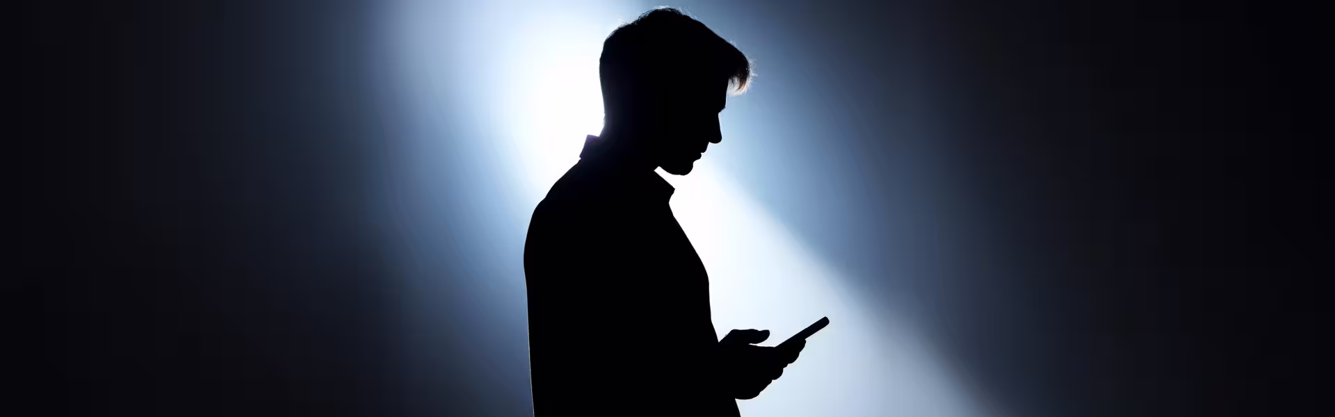 Silhouette of a person in profile looking down at a smartphone, backlit by a soft blue light that creates a high-contrast outline against a dark background