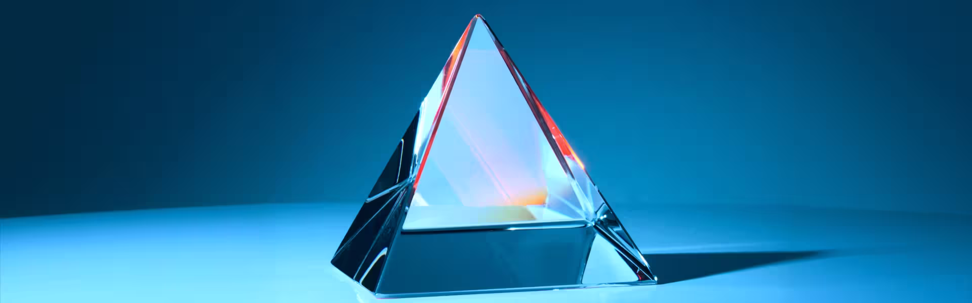 Glass pyramid prism on a blue surface, refracting light with subtle red and cyan highlights against a clean blue background, creating a sharp, geometric, and reflective composition