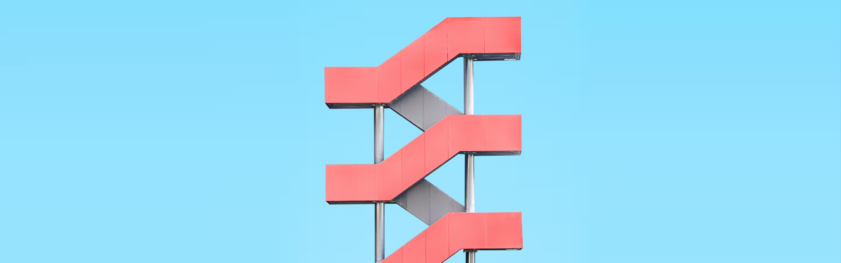 A modern, abstract staircase with red steps and gray supports against a bright blue sky, creating a striking architectural element.