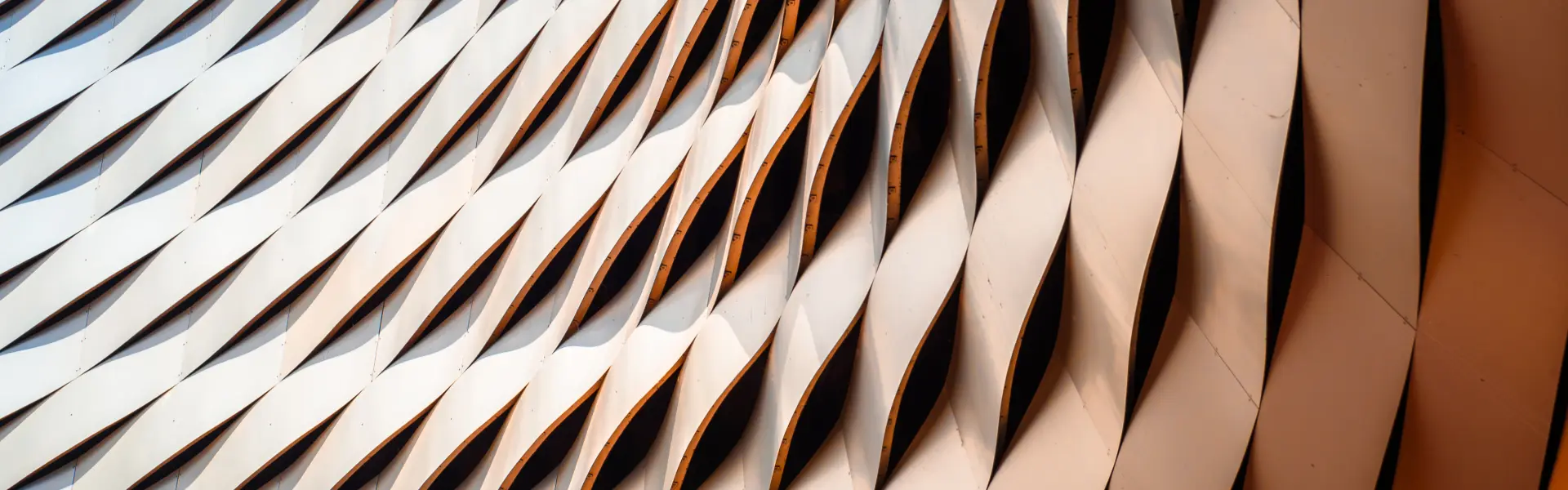 Abstract architectural facade featuring a repeating wave-like pattern of twisted metal panels. The structure creates a dynamic play of light and shadow across the surface.