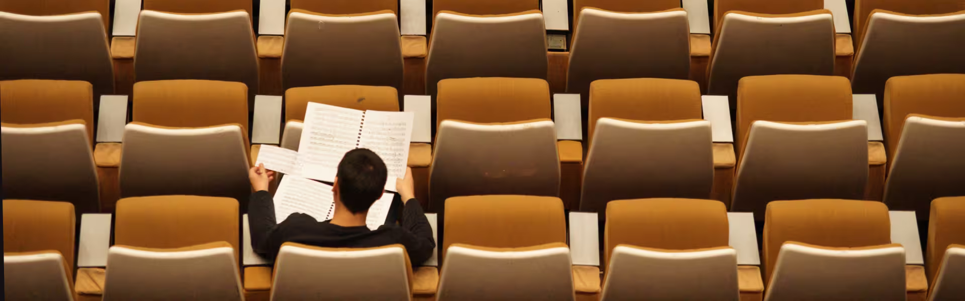 Student sitting alone in an empty lecture hall, reviewing open music sheets in a notebook, symbolizing focused learning and study in education