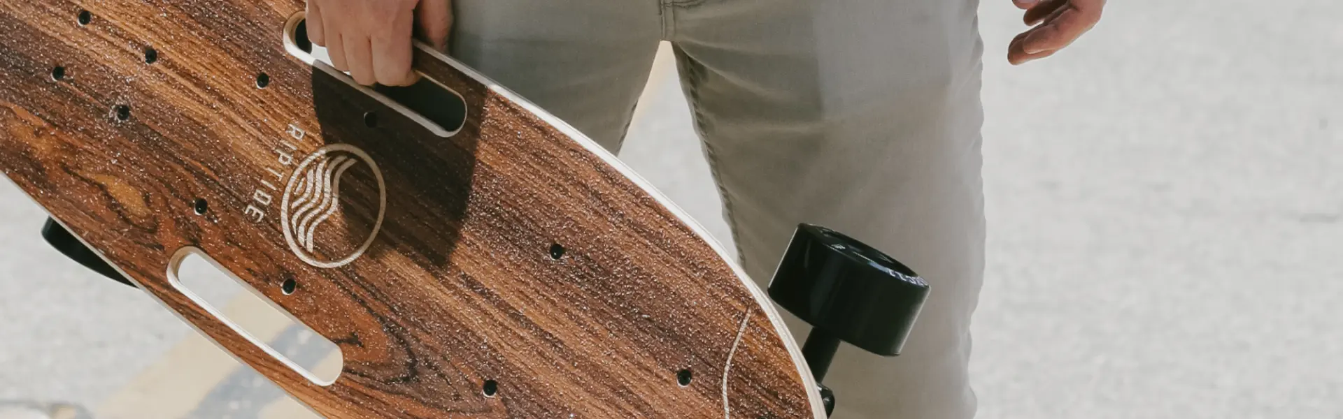 Close-up of a person holding a wooden longboard with black wheels and built-in hand grips. The deck features a wavy line design and the branding "RIPTIDE" etched into the wood.