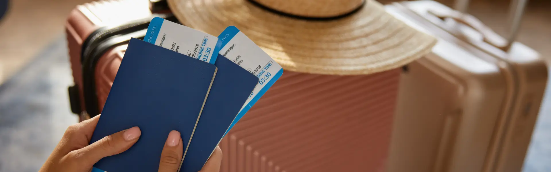 Close-up of a person holding two blue passports with boarding passes, with pink suitcases and a straw hat in the background, suggesting travel or vacation.