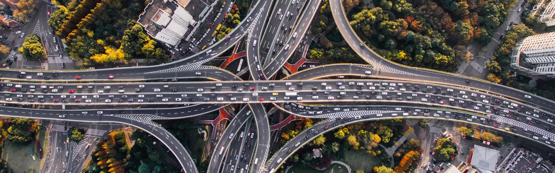 Aerial view of a busy multi-level highway interchange with heavy traffic, showcasing numerous lanes, ramps, and overpasses surrounded by trees and urban buildings.