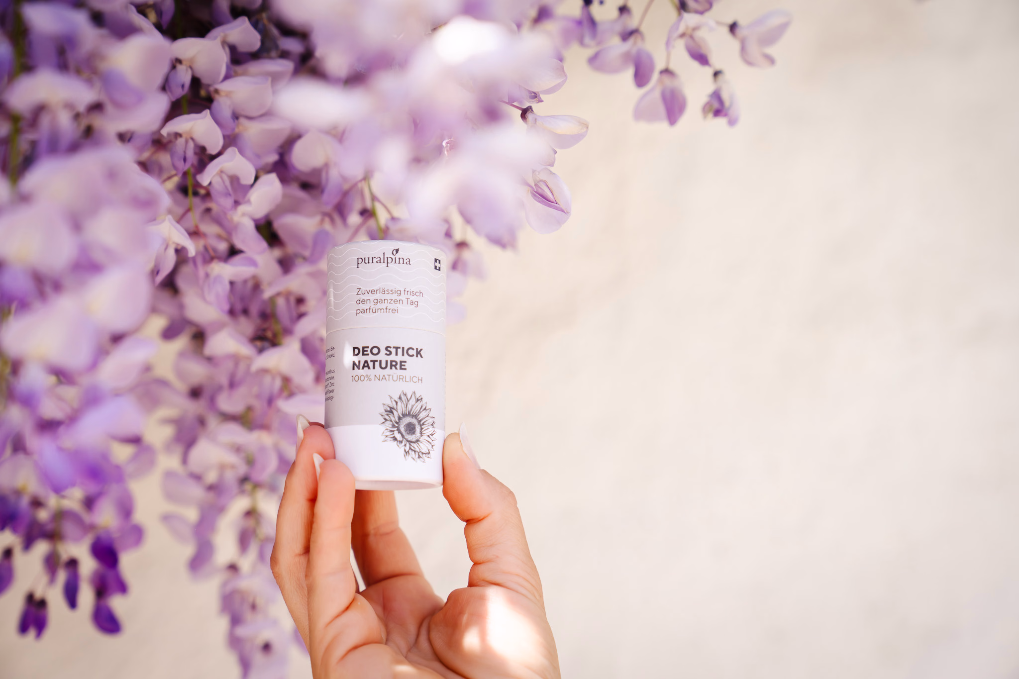 Hand hält Puralpina Deo Stick Nature vor blühendem Blauregen an heller Fassade – Foto von Chantal Reichenbach für deeds