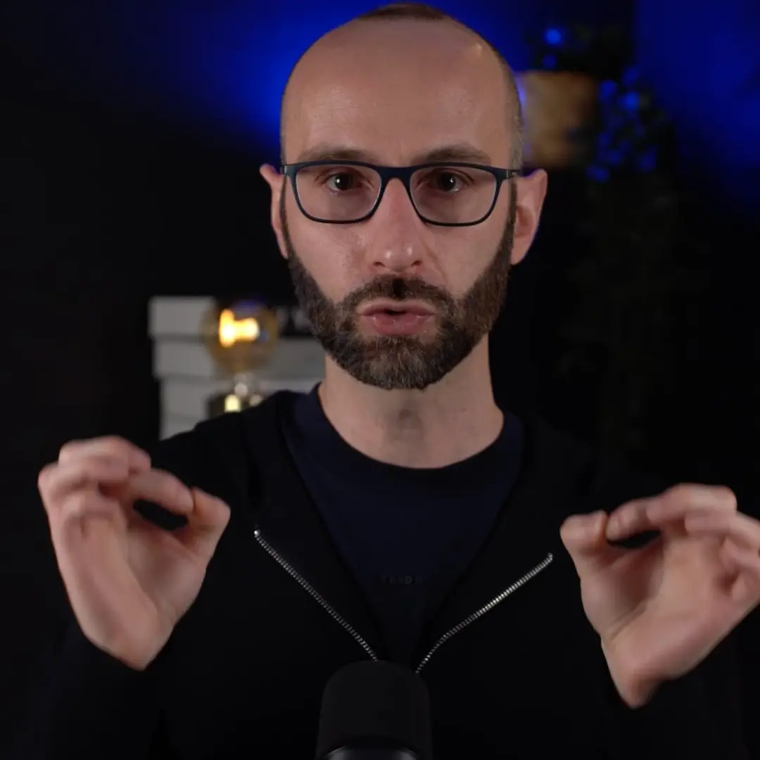 Man with glasses and beard speaking with hands raised in front of a dark background with a blurred light.