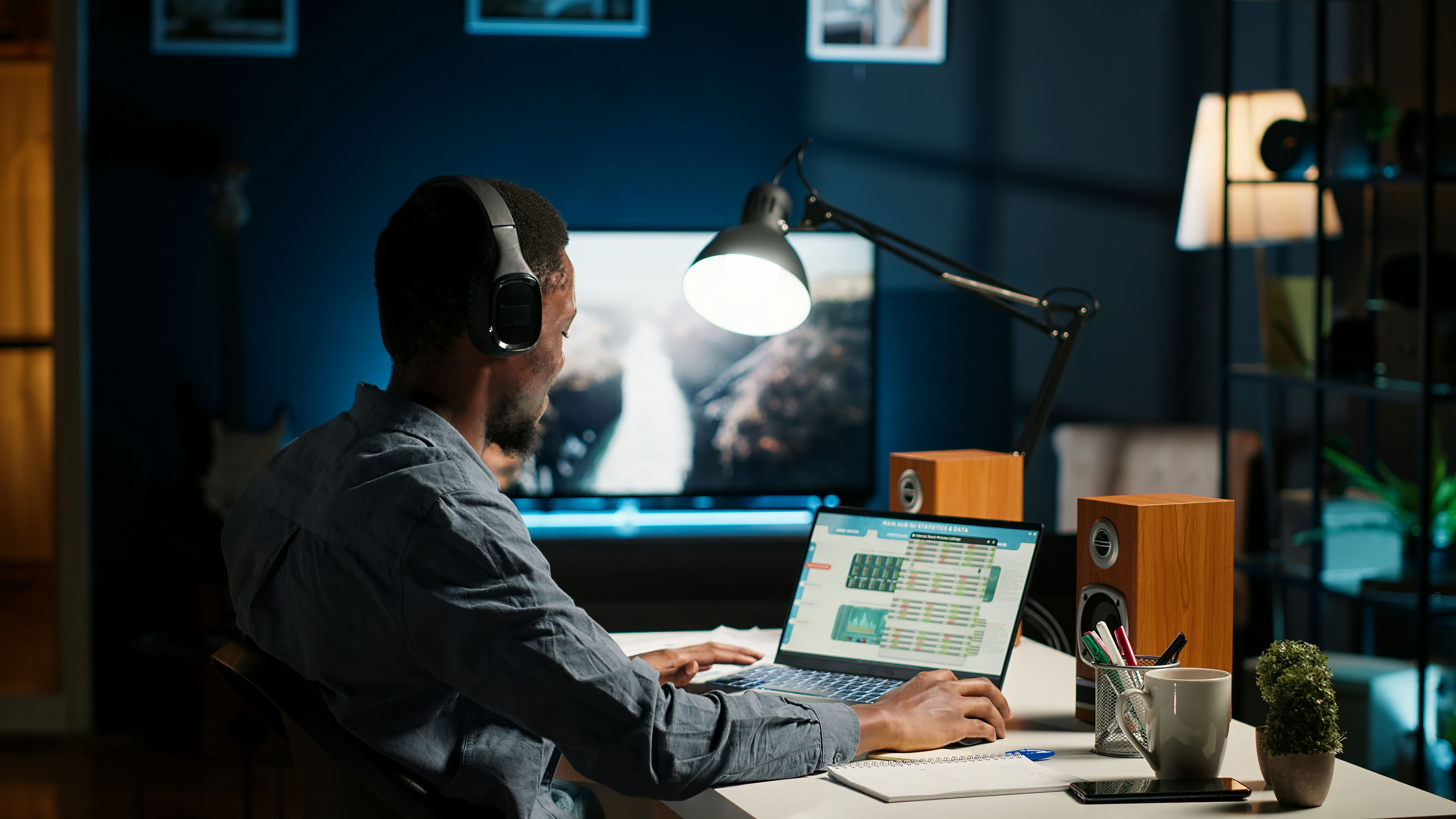 A man wearing headphones sits at a home desk at night, typing on a laptop displaying a colorful data dashboard, with a desk lamp, wooden speakers, and a large monitor visible in the background