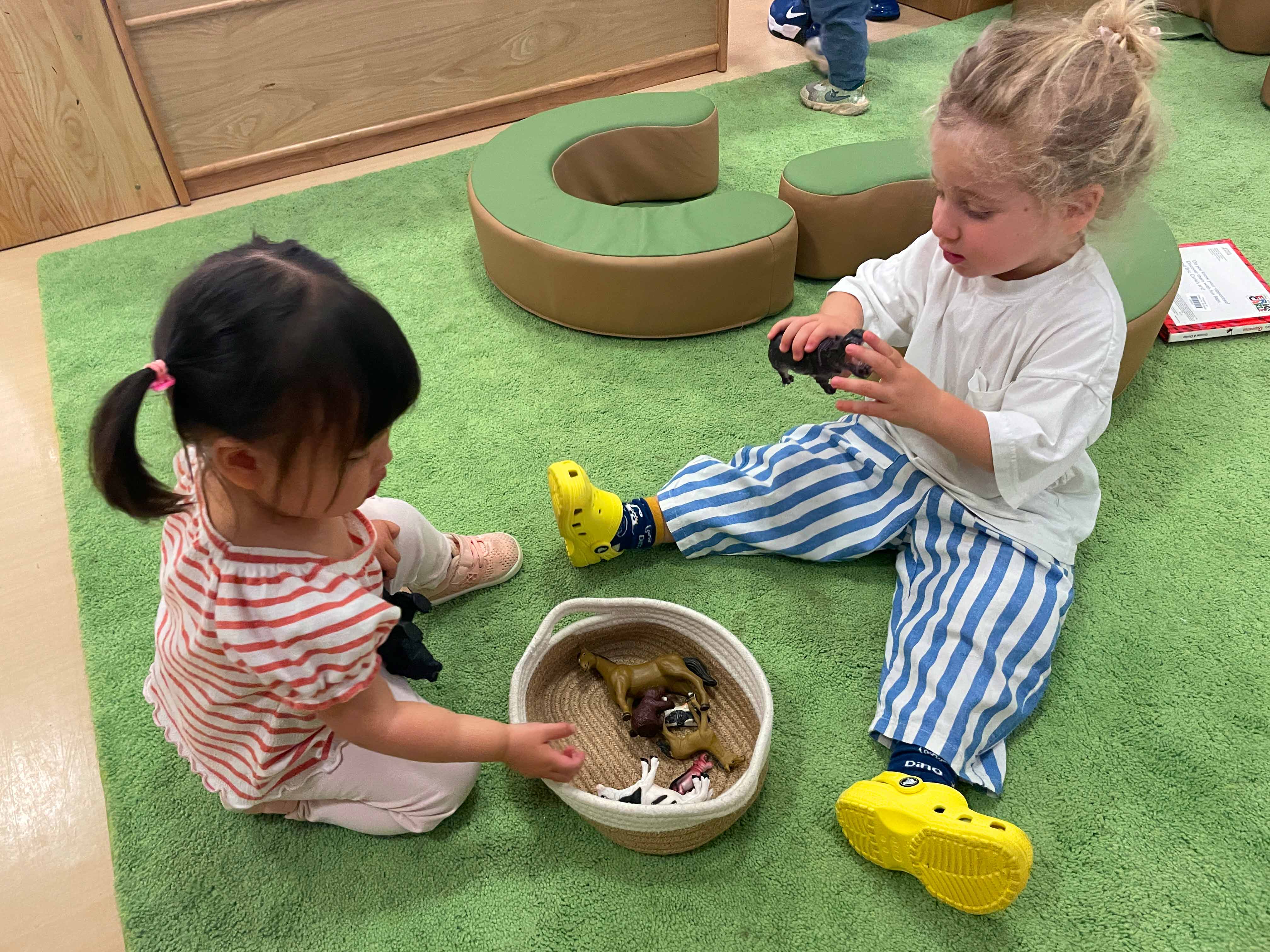 Two students seated together on the classroom carpet are playing together and sharing animal toy figures.