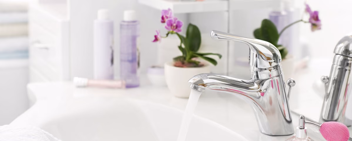 Modern chrome bathroom faucet with running water and orchid plant on white sink counter.
