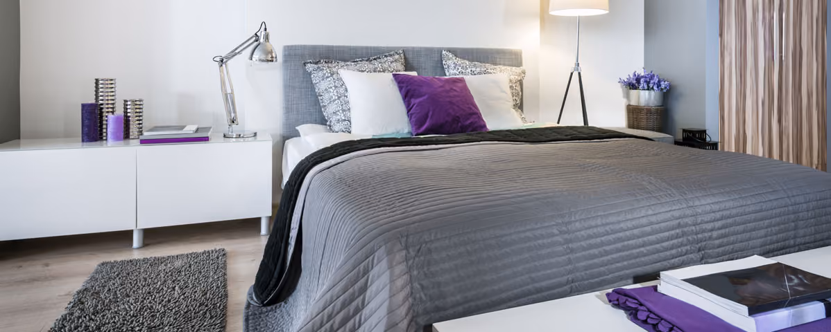Modern bedroom with gray bedspread, purple and white pillows, white furniture, and a lamp with flowers.