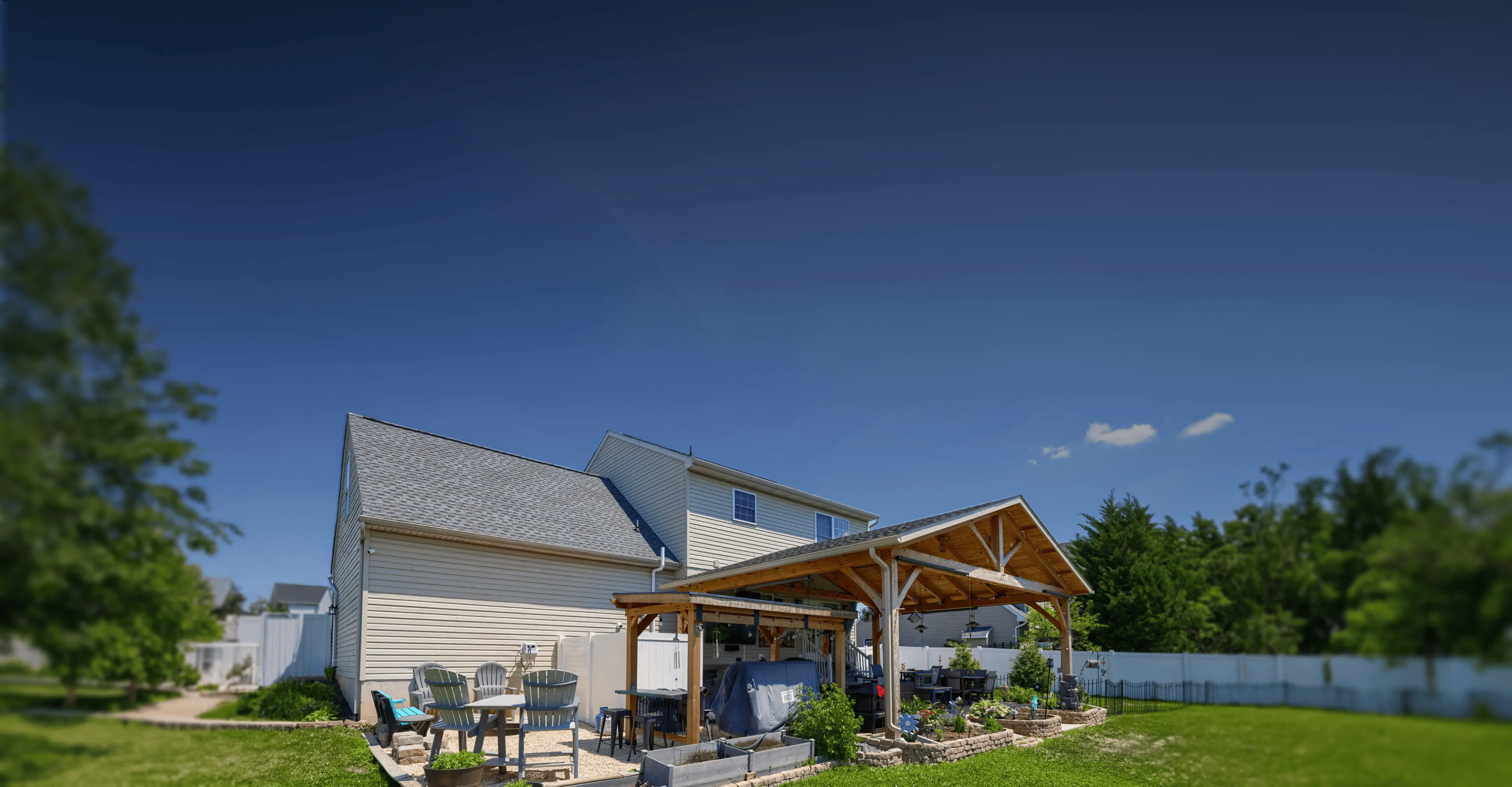 A backyard with green grass, a wooden pergola over a patio area, outdoor seating, plants, and a white house in the background under a clear blue sky.