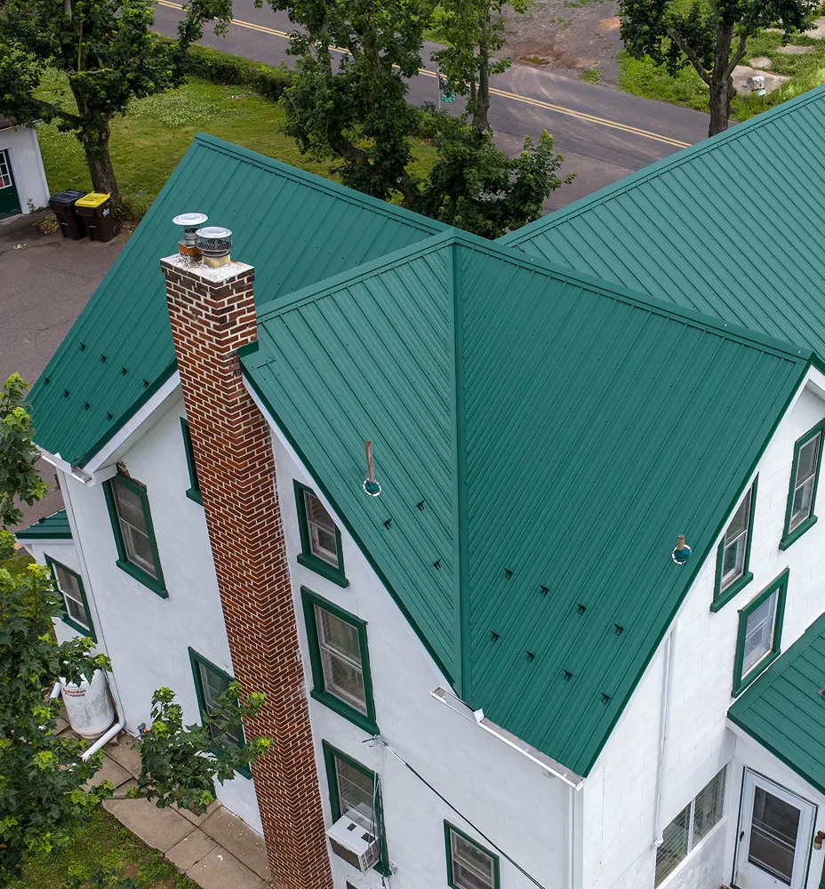 Aerial view of a white house with a green metal roof, brick chimney, and multiple windows, surrounded by trees and a nearby road.