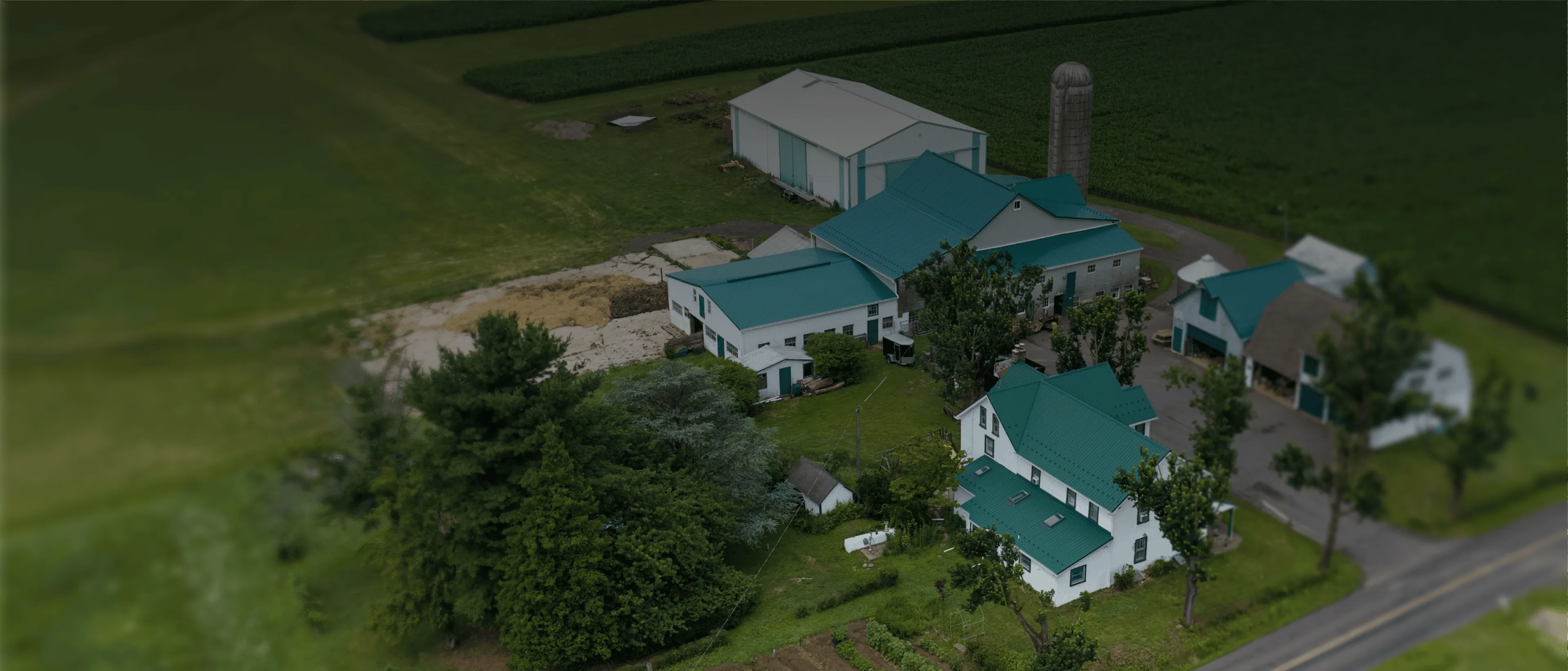 Aerial view of a farmstead with white buildings, tasteful new green roofs, a silo, and surrounding farmland.