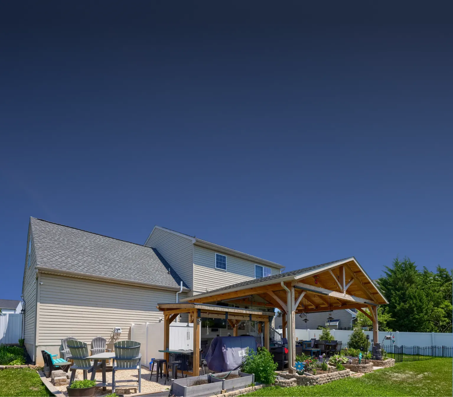 A backyard with green grass, a wooden pergola over a patio area, outdoor seating, plants, and a white house in the background under a clear blue sky.