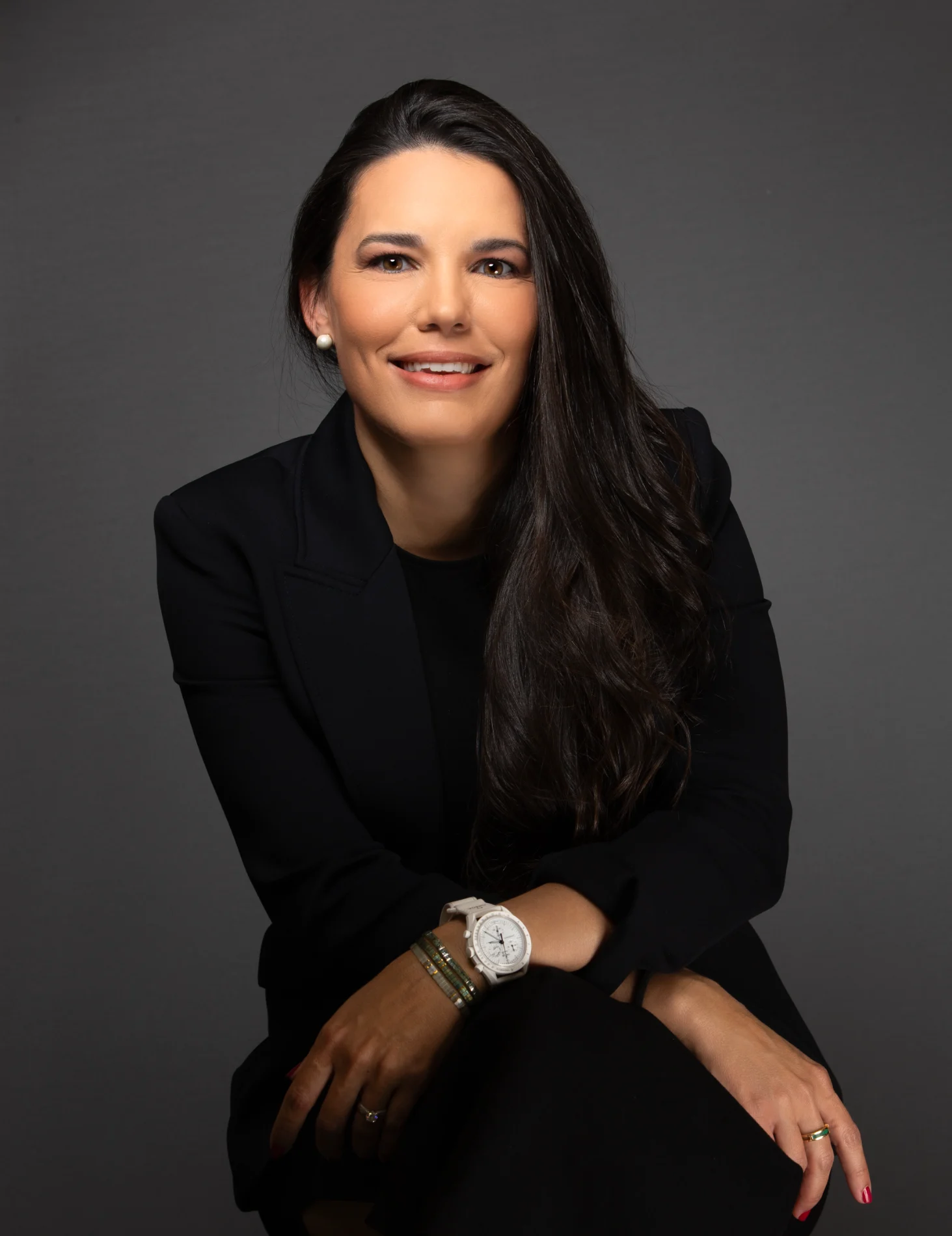 Smiling woman with long dark hair, black blazer, and accessories sitting against a gray background.